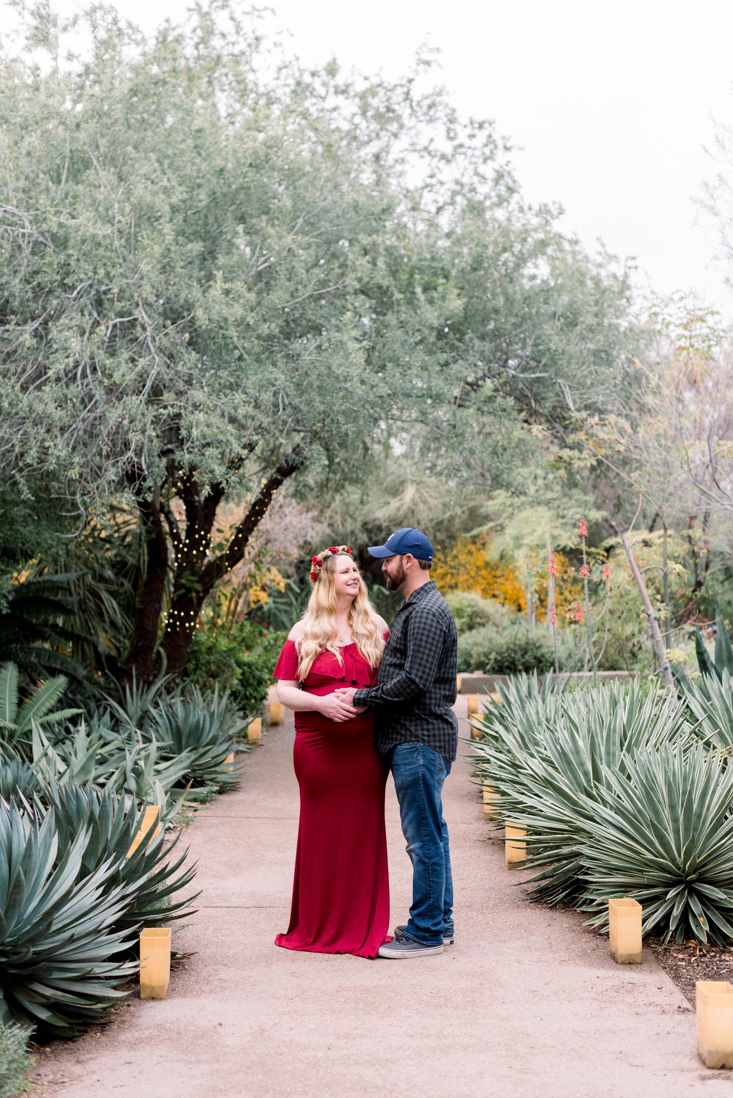 Romantic maternity photo in a red dress along a garden path lined with desert plants and string lights.