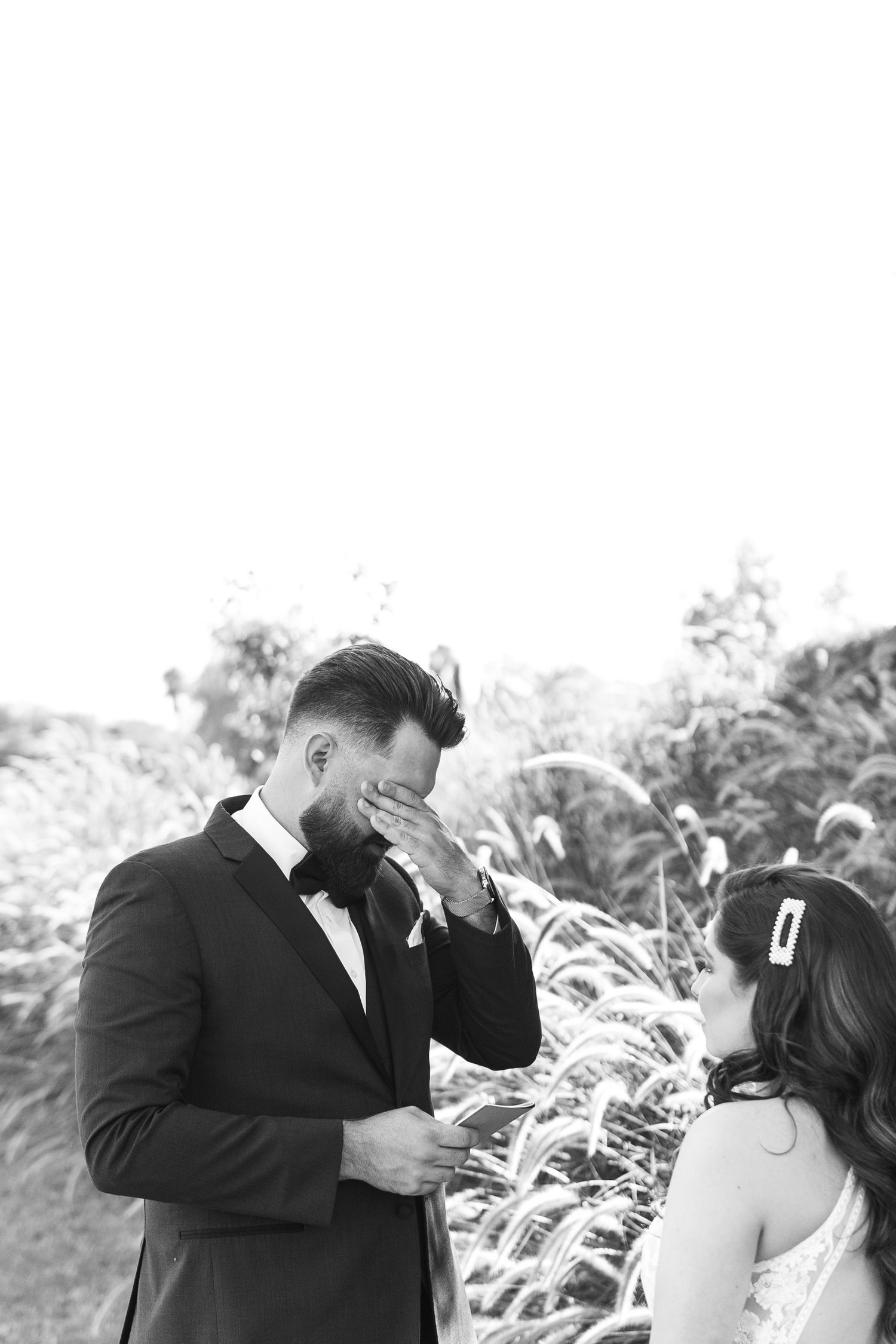 An emotional black and white moment showing a groom in a tuxedo wiping tears during an outdoor wedding ceremony.
