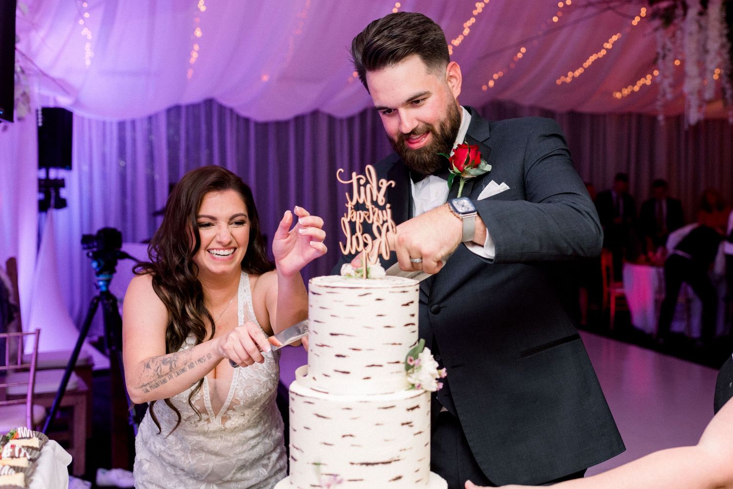 Newlyweds share a playful moment while cutting their tall white wedding cake.
