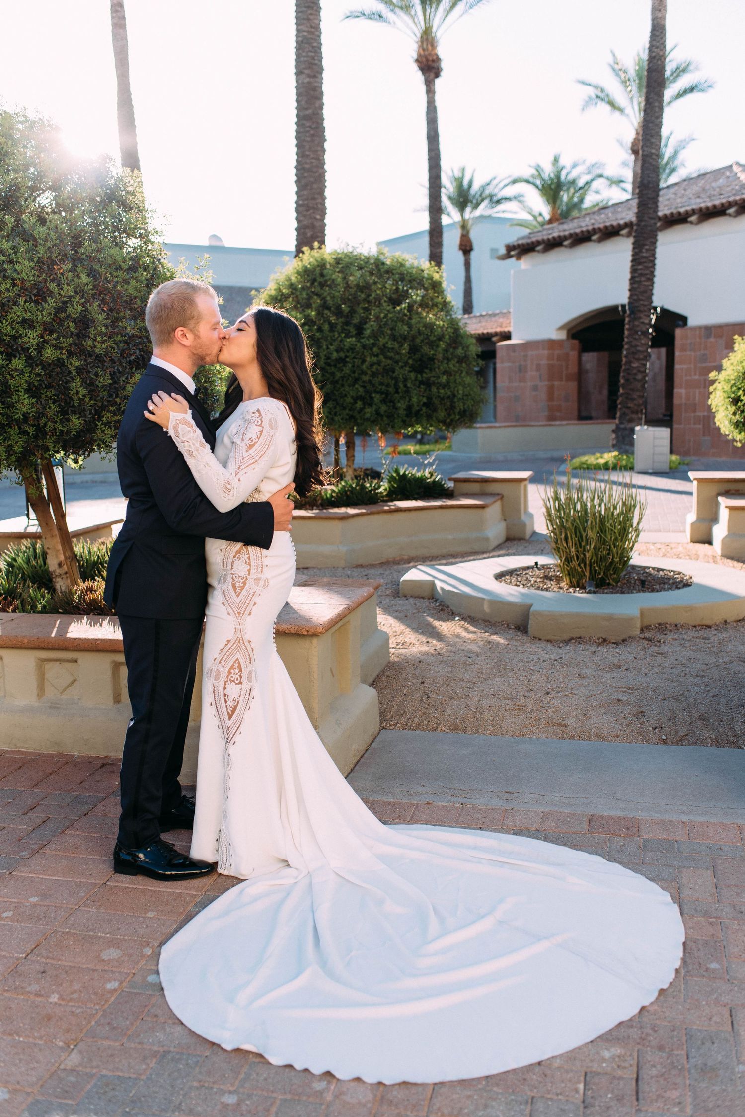 Wedding couple embracing in courtyard with palm trees and dramatic train in foreground.