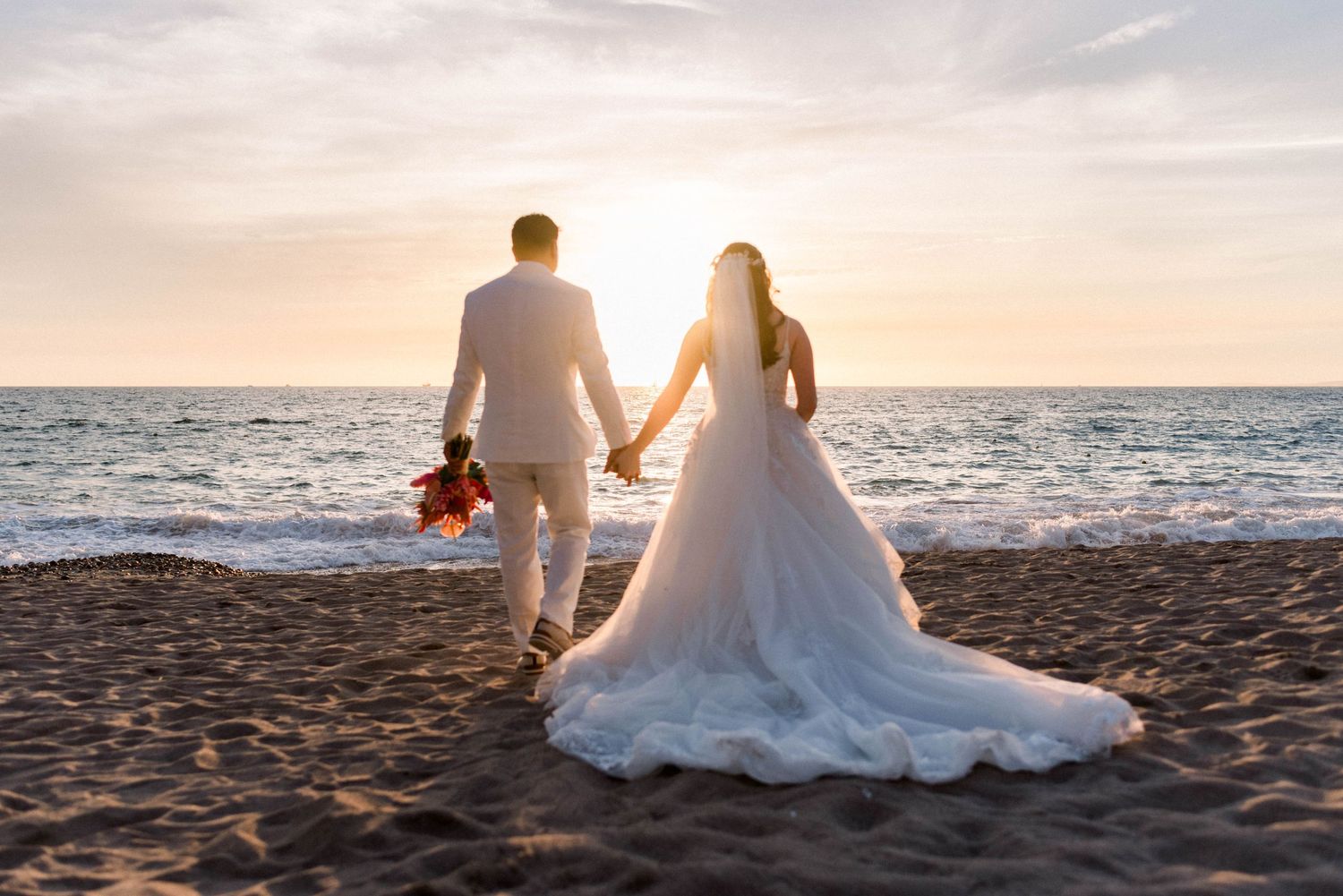 Couple walking on sandy beach at sunset in wedding attire with long white dress trailing behind.