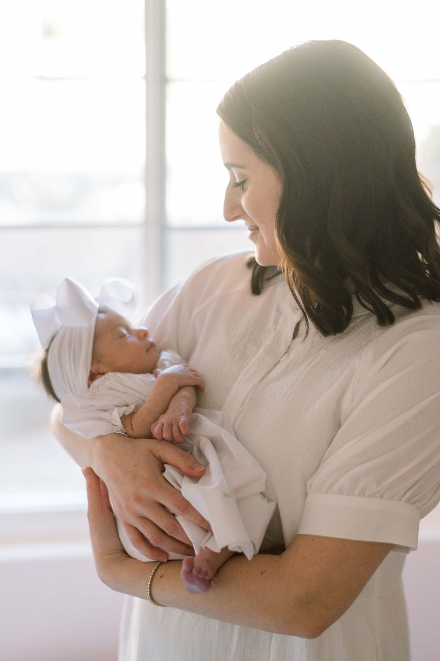 Mother in white dress holds sleeping newborn swaddled in white blanket near window.