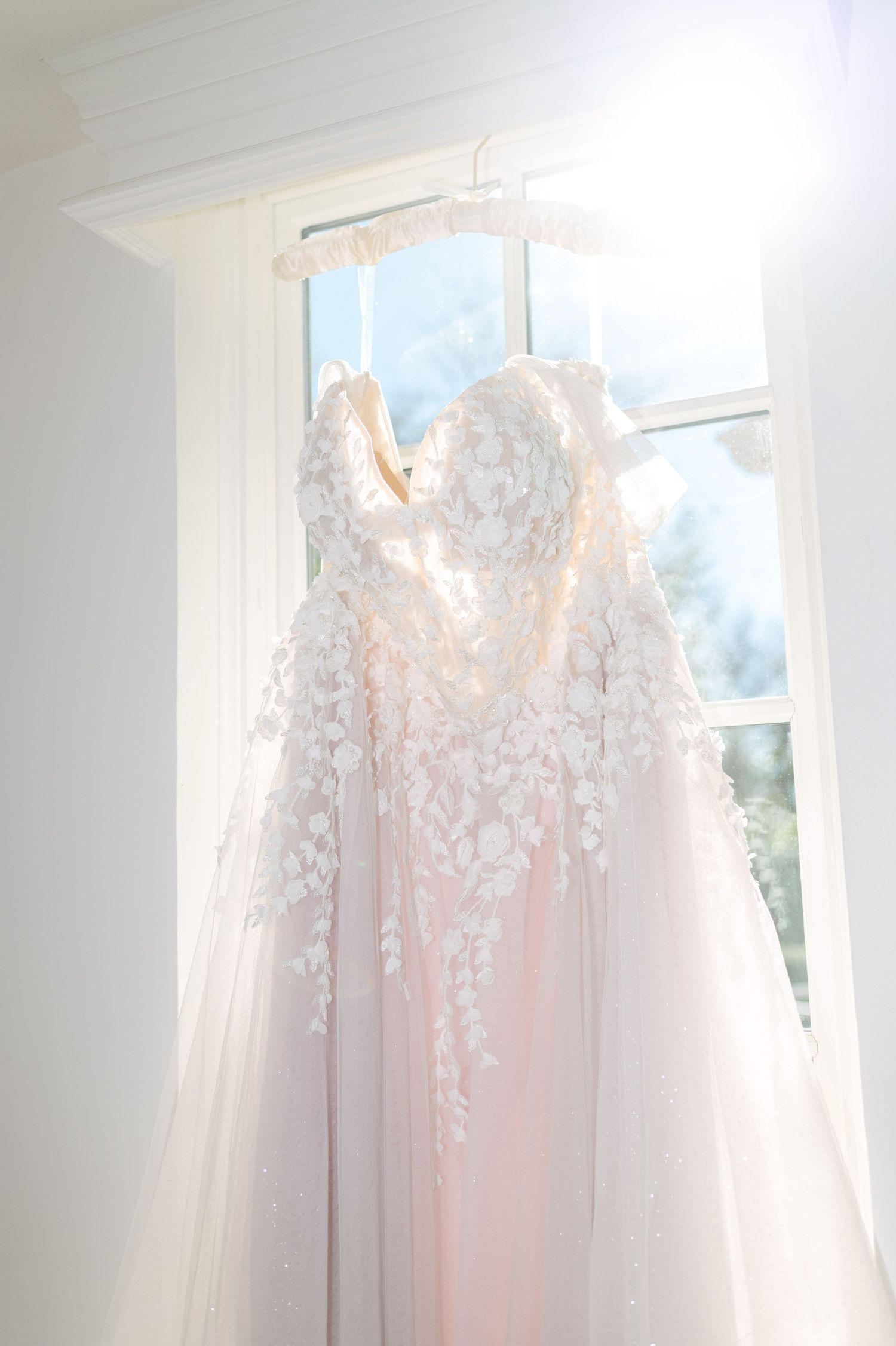 Delicate white wedding dresses hanging in sunlit window.