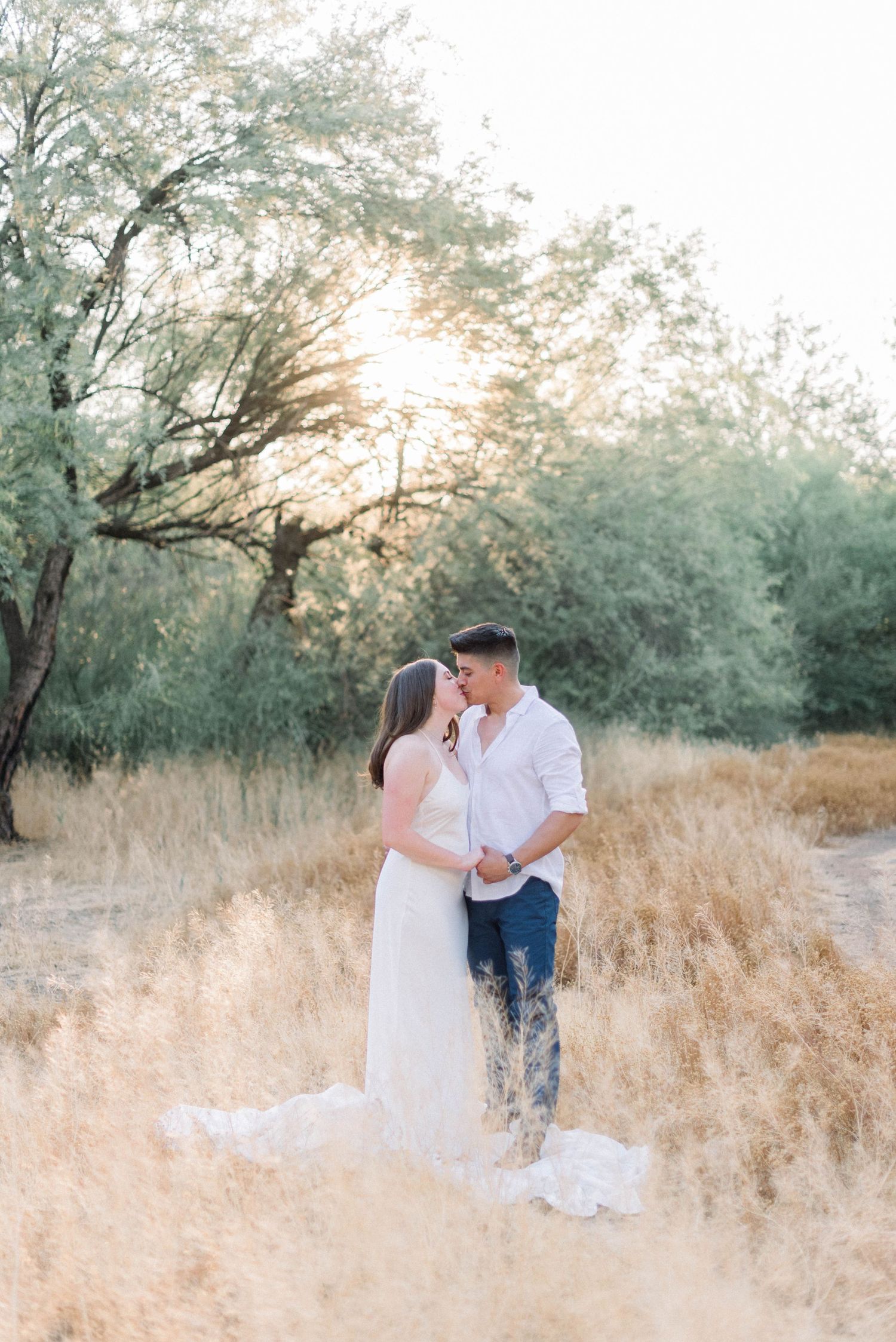 A couple in white attire shares romantic moments in a sunlit field with tall grass and trees at golden hour.