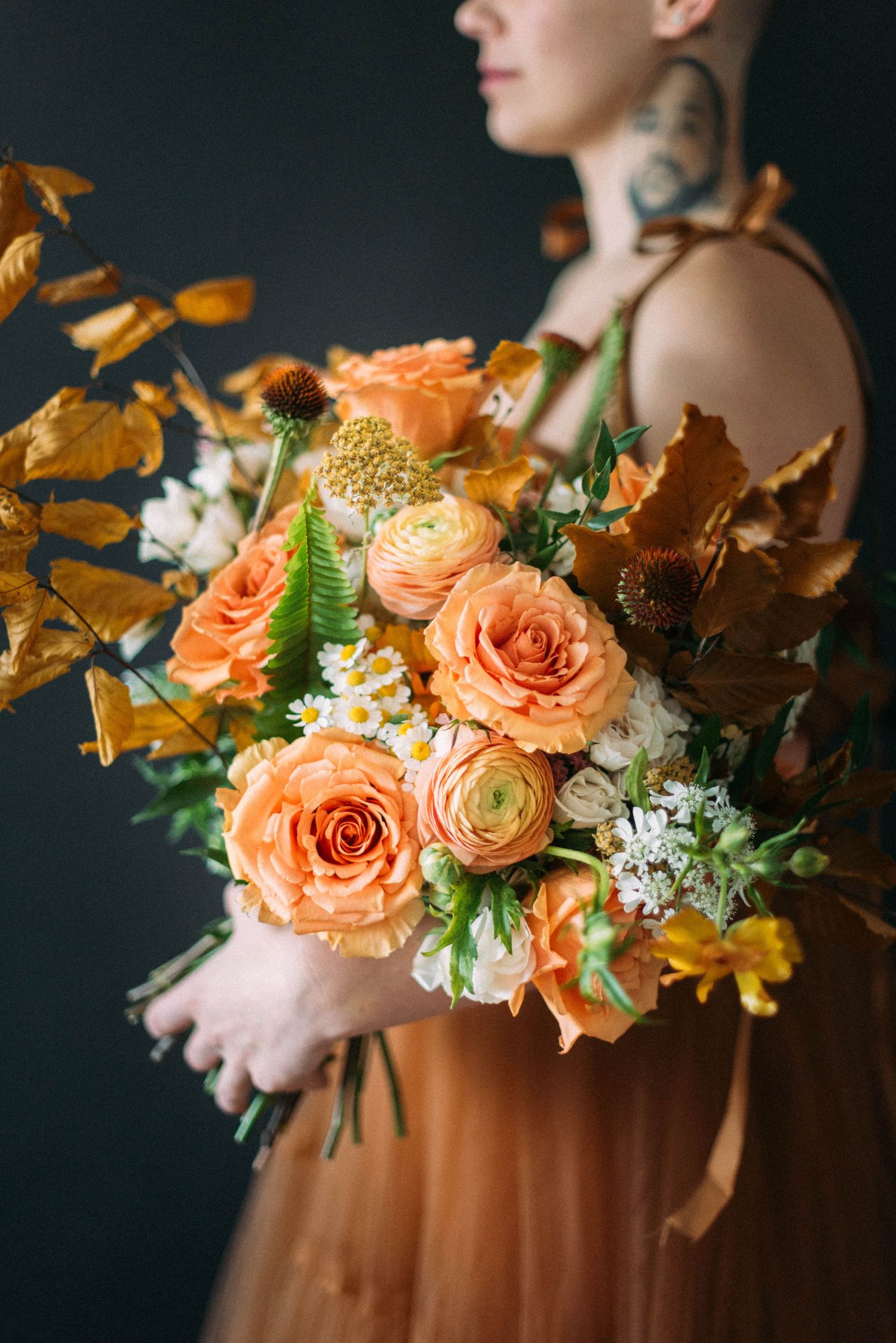 A moody floral arrangement featuring peach roses and autumn leaves held against a dark background.