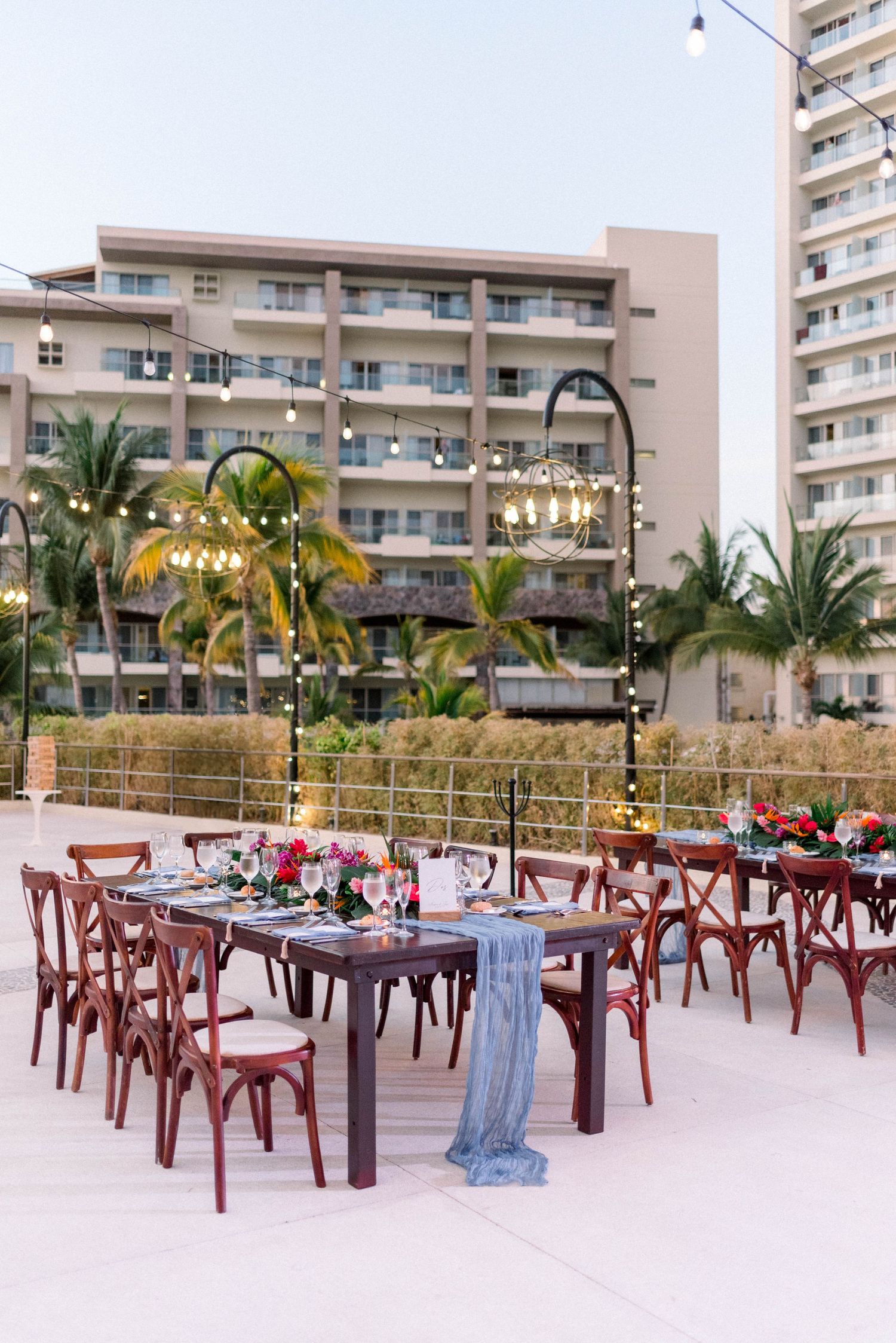 Tropical wedding reception setup with wooden cross-back chairs and blue table runners.
