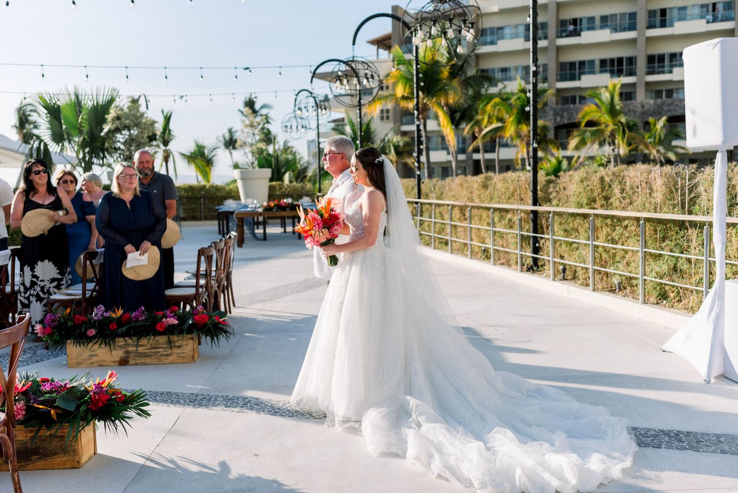 Bride in white gown holding colorful bouquet during outdoor beachfront ceremony.