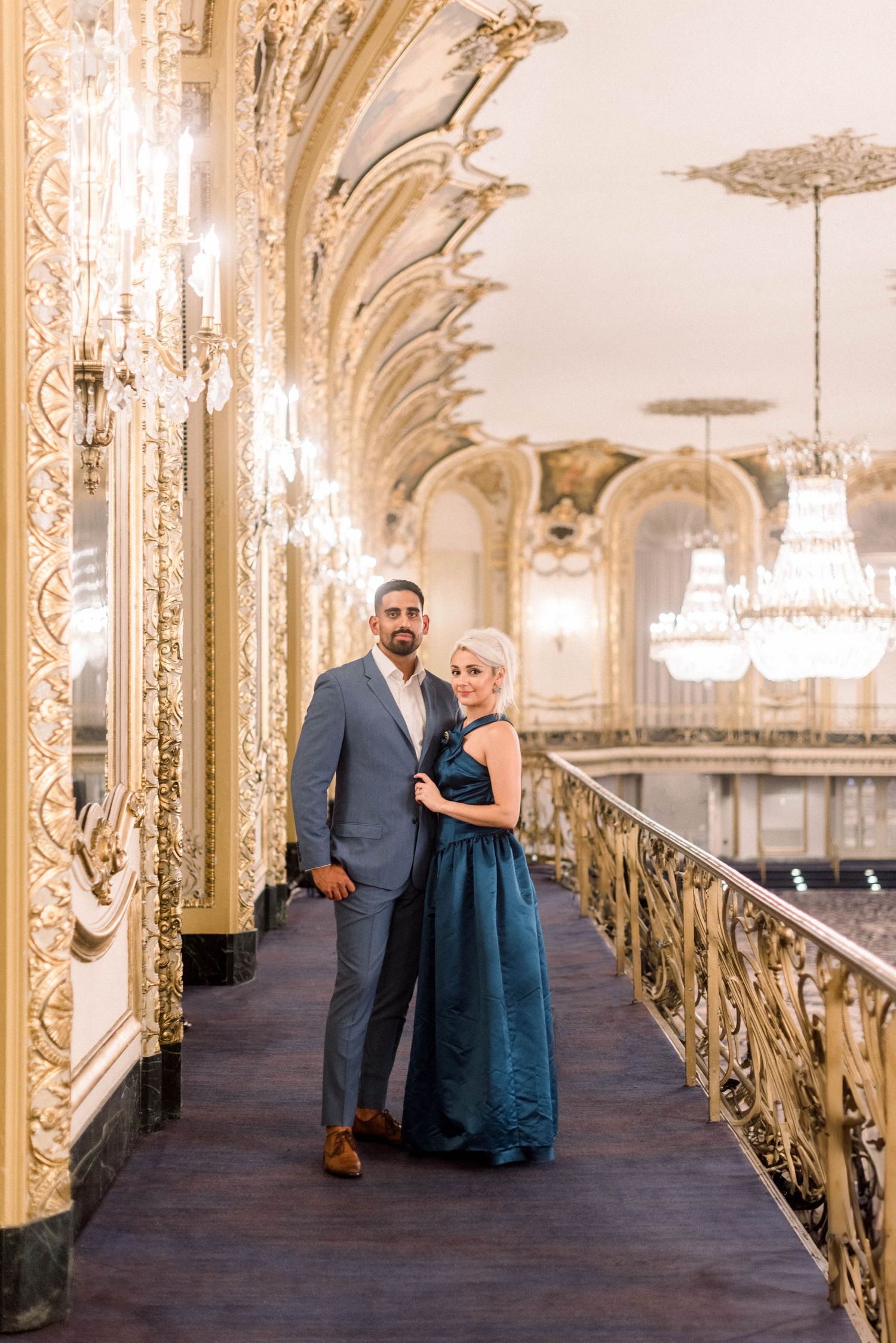 A couple poses elegantly in formal wear along an ornate gilded hallway with crystal chandeliers.