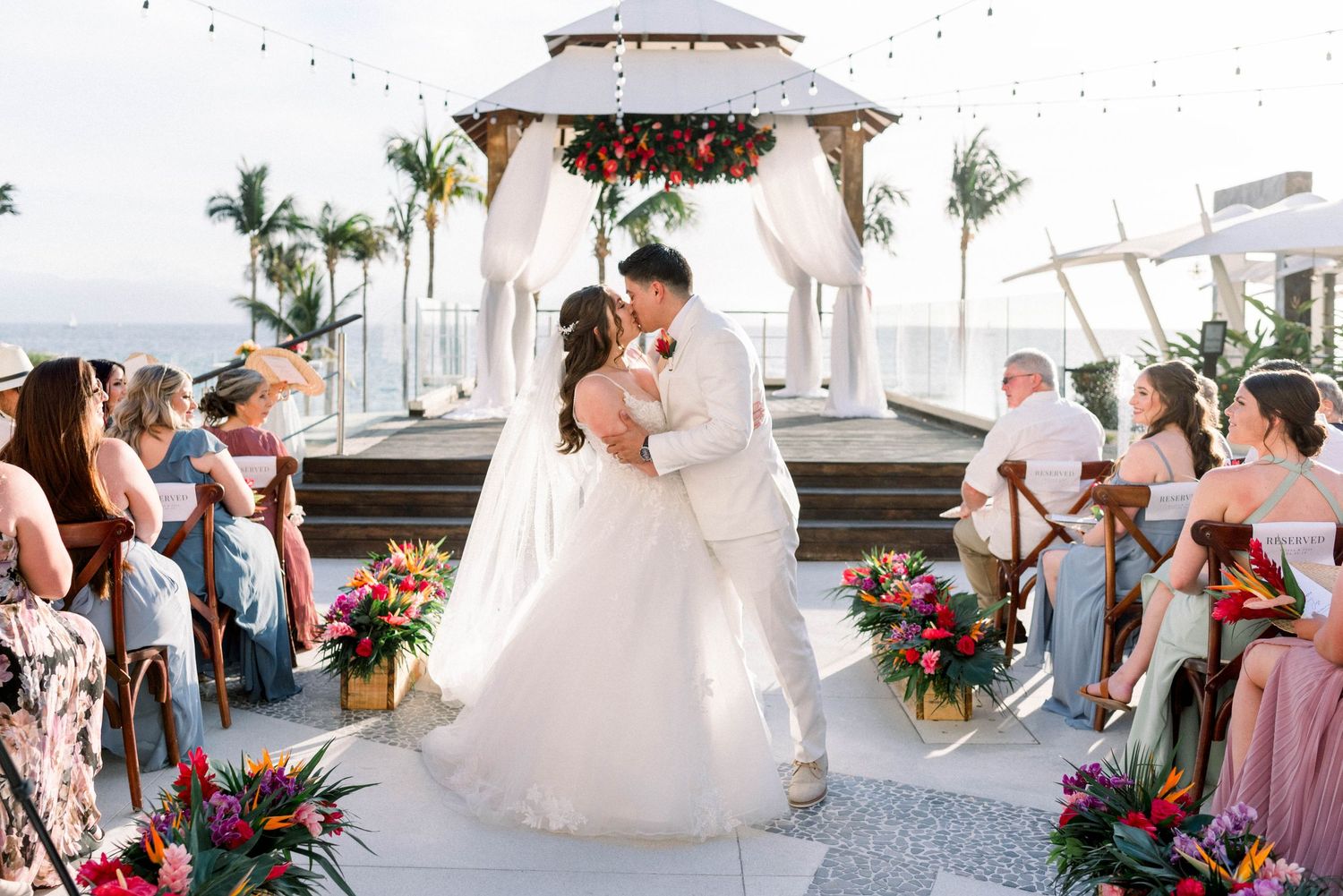 A wedding ceremony on a decorated white gazebo with tropical palm trees and flower arrangements.