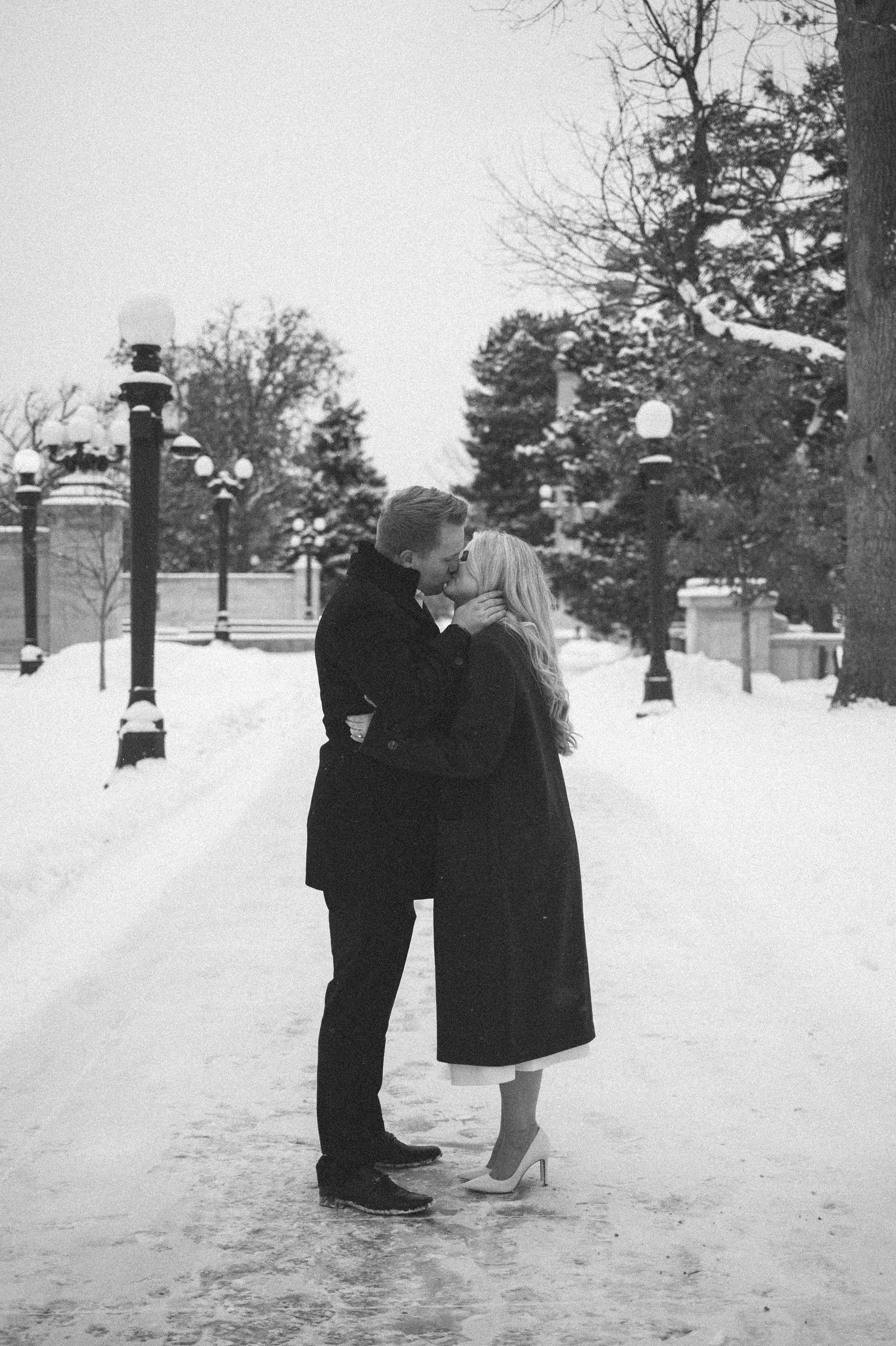 A romantic winter embrace between a couple on a snowy path lined with vintage lampposts.