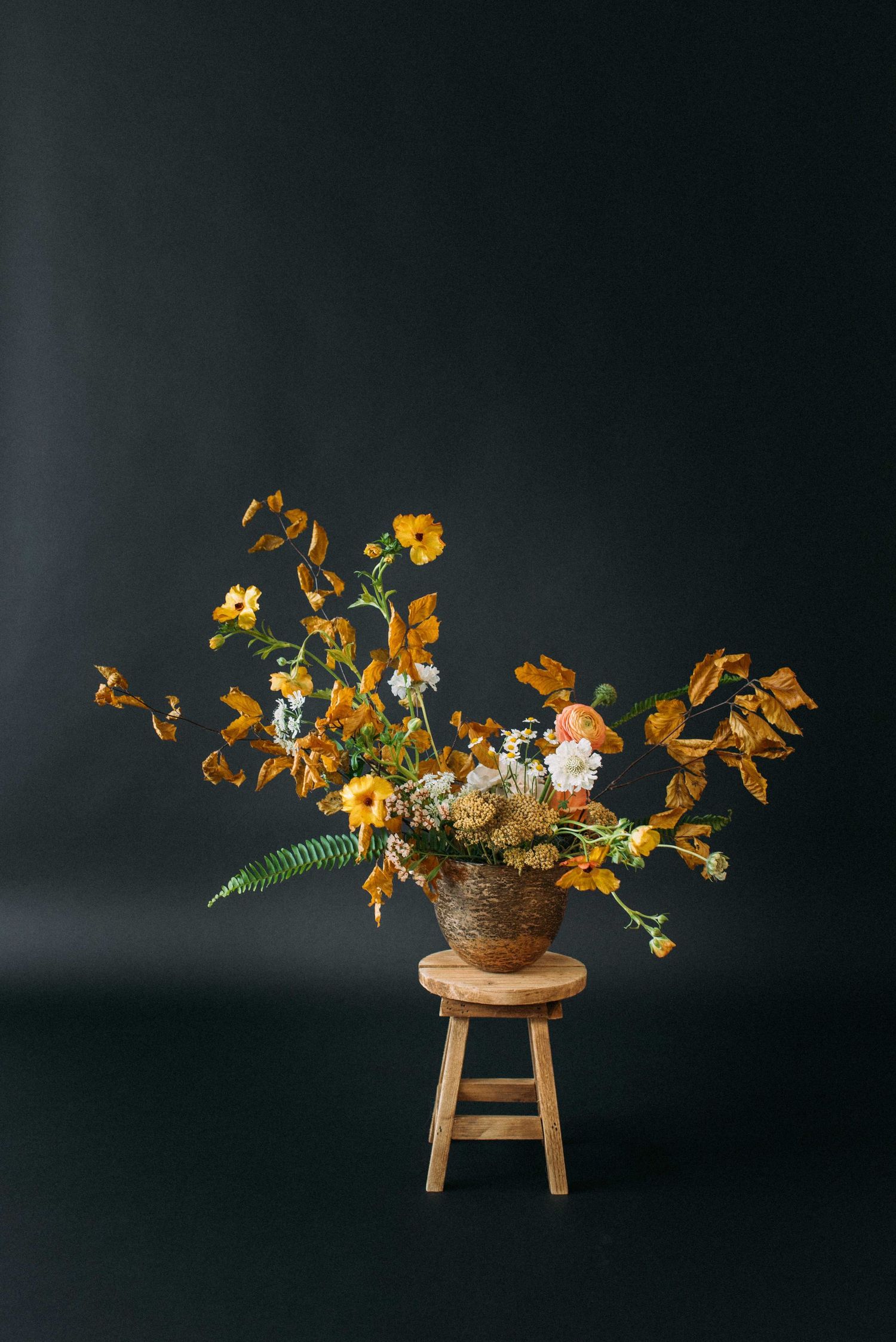 Dramatic floral arrangement with autumn-colored blooms displayed on wooden stool against dark background.