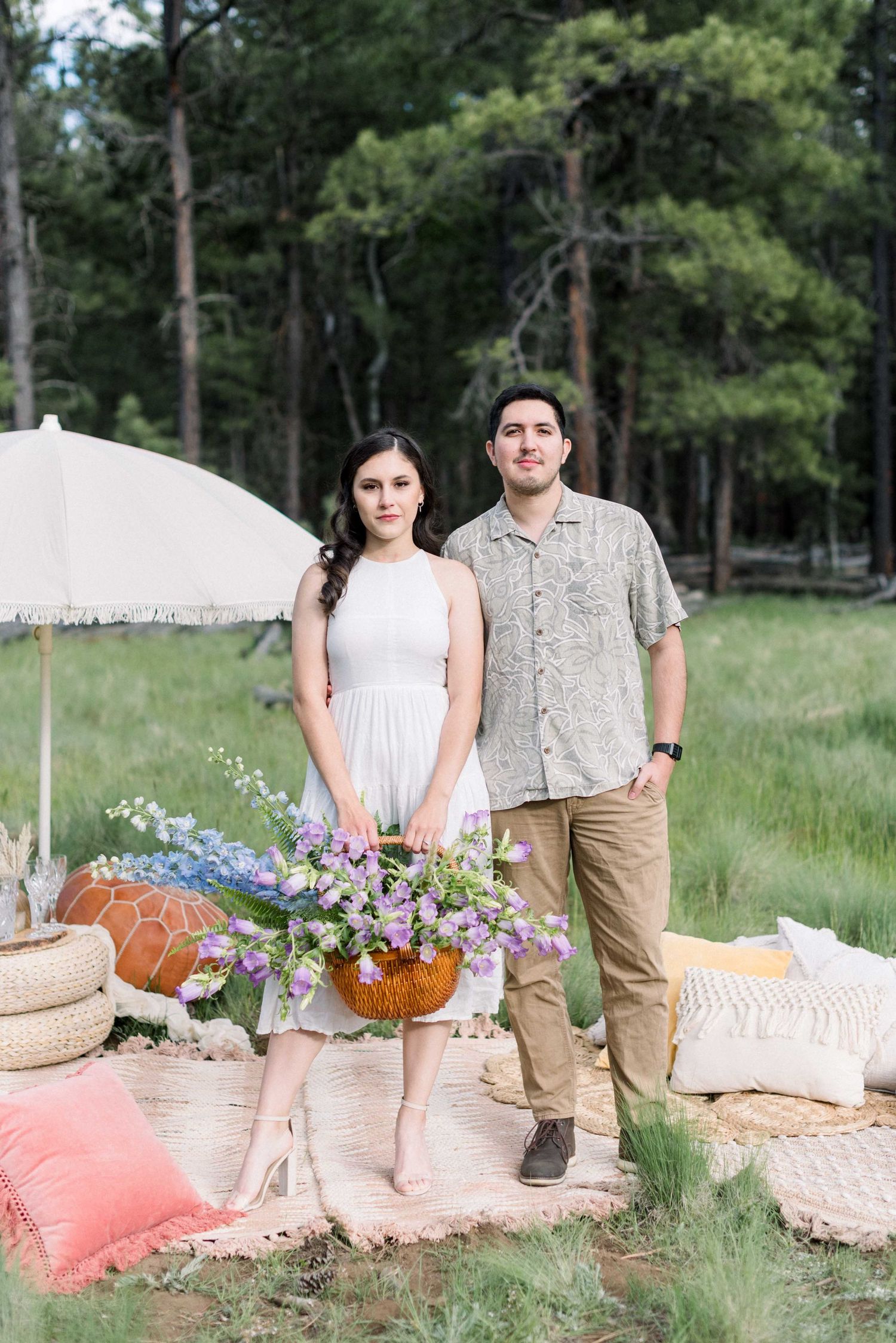 A romantic picnic setup in a forest with white umbrella, pillows and wildflower arrangements.