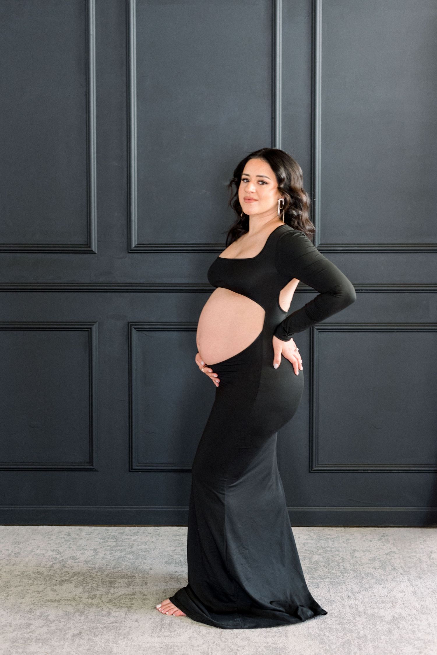 Stylish maternity photo in black dress against dark paneled wall backdrop.