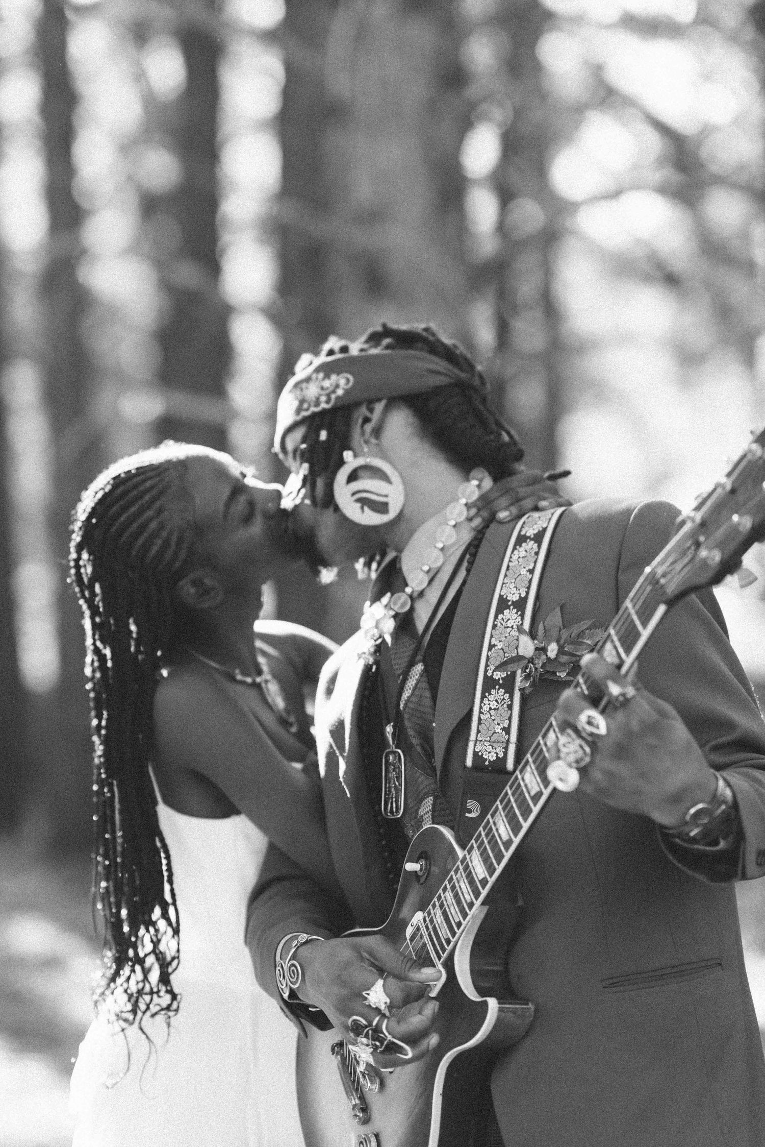 A black and white candid photo of newlyweds laughing together with an electric guitar in a forest.