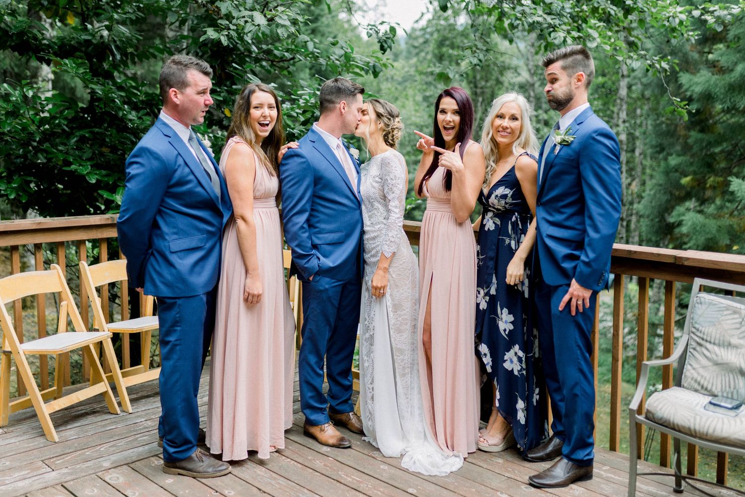 Wedding party in coordinated blue suits and pink dresses poses on wooden deck.