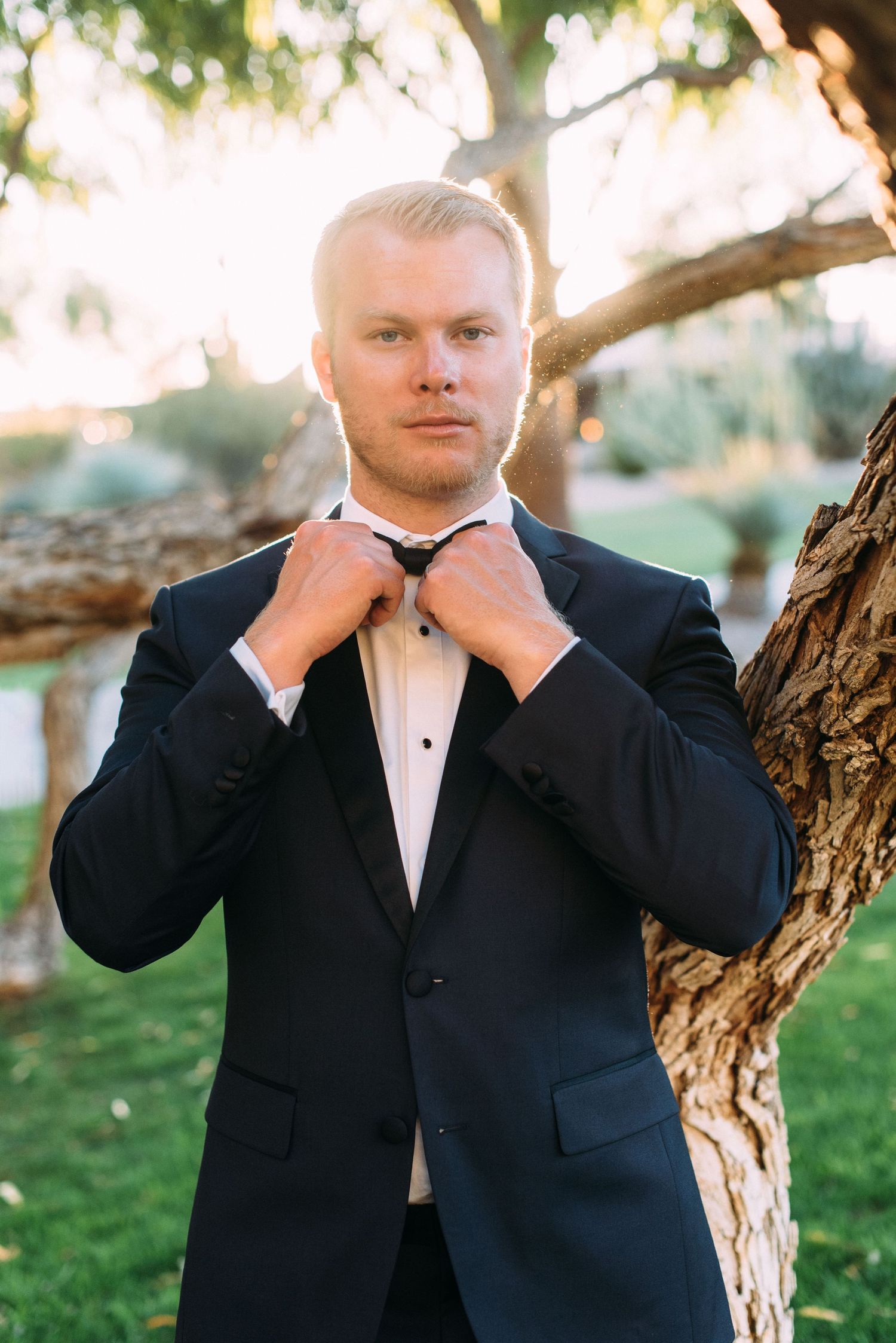 A groom in a classic black tuxedo adjusts his bowtie while standing near a tree.