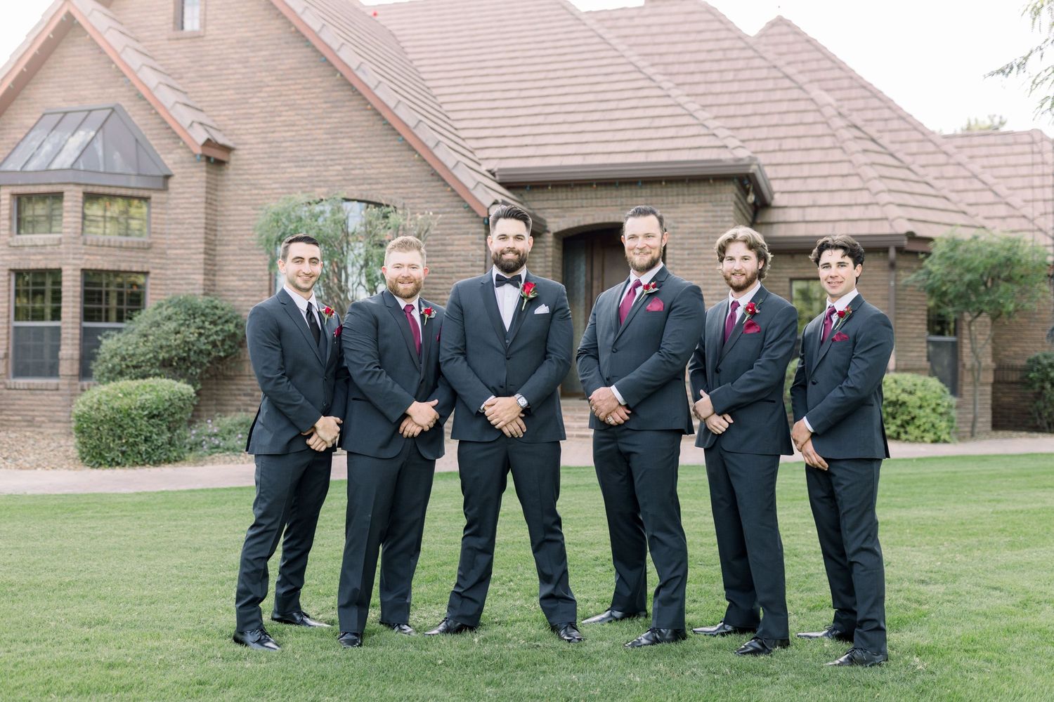 Groomsmen in dark suits and red ties pose formally on lawn in front of venue.