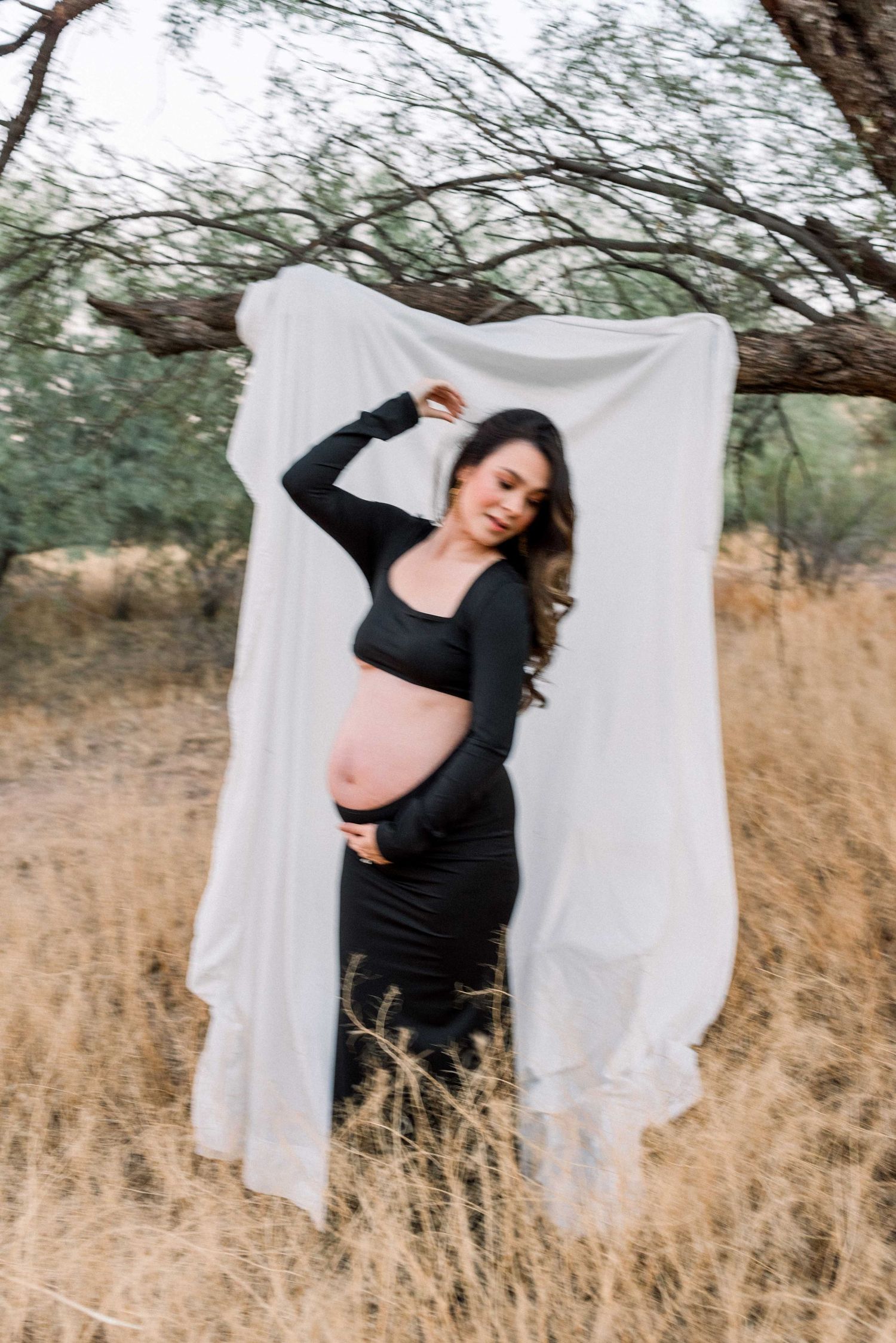 Candid maternity portrait outdoors with flowing white fabric backdrop and natural desert landscape.
