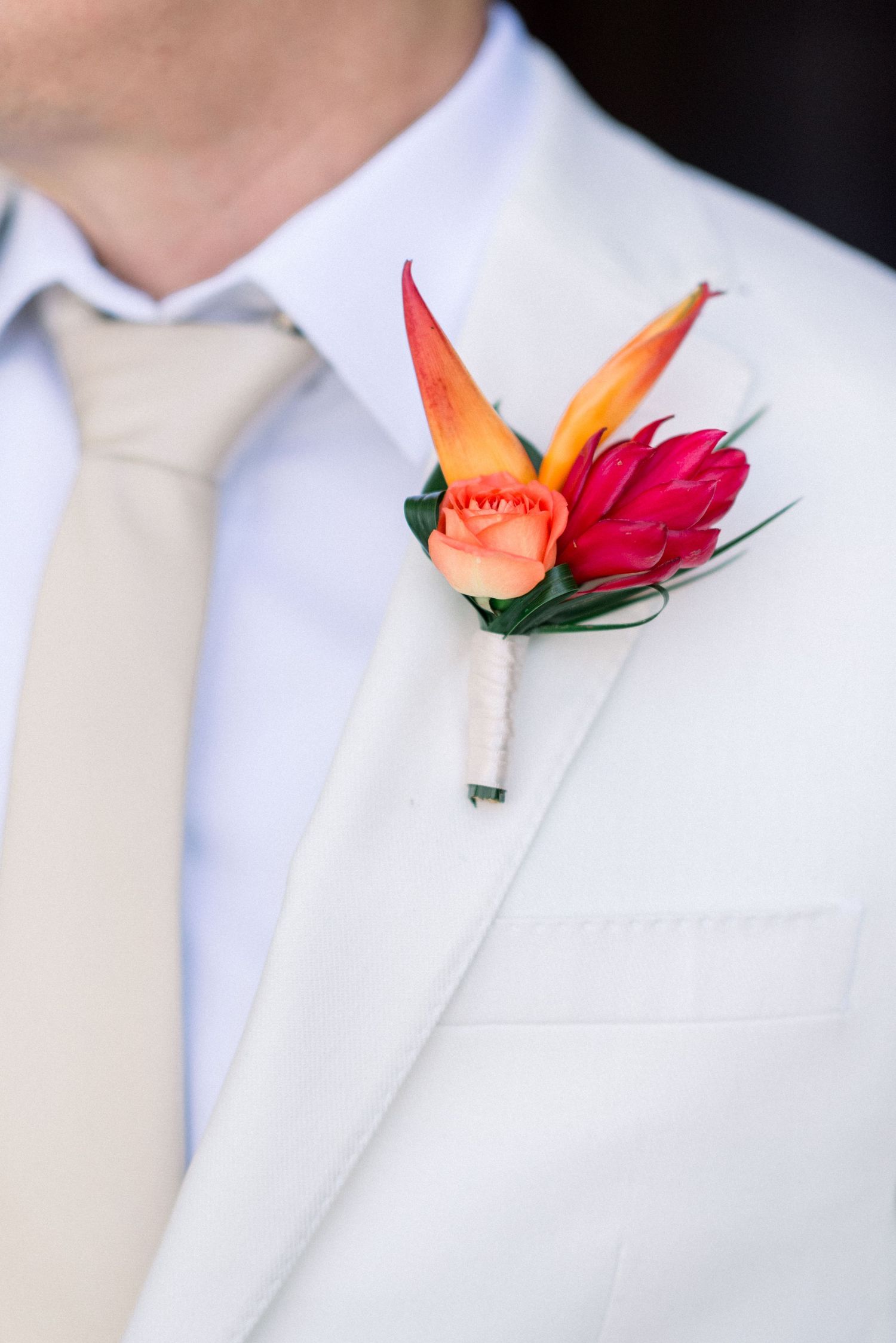 Elegant white boutonniere with orange bird of paradise and red accent flower on light suit jacket.