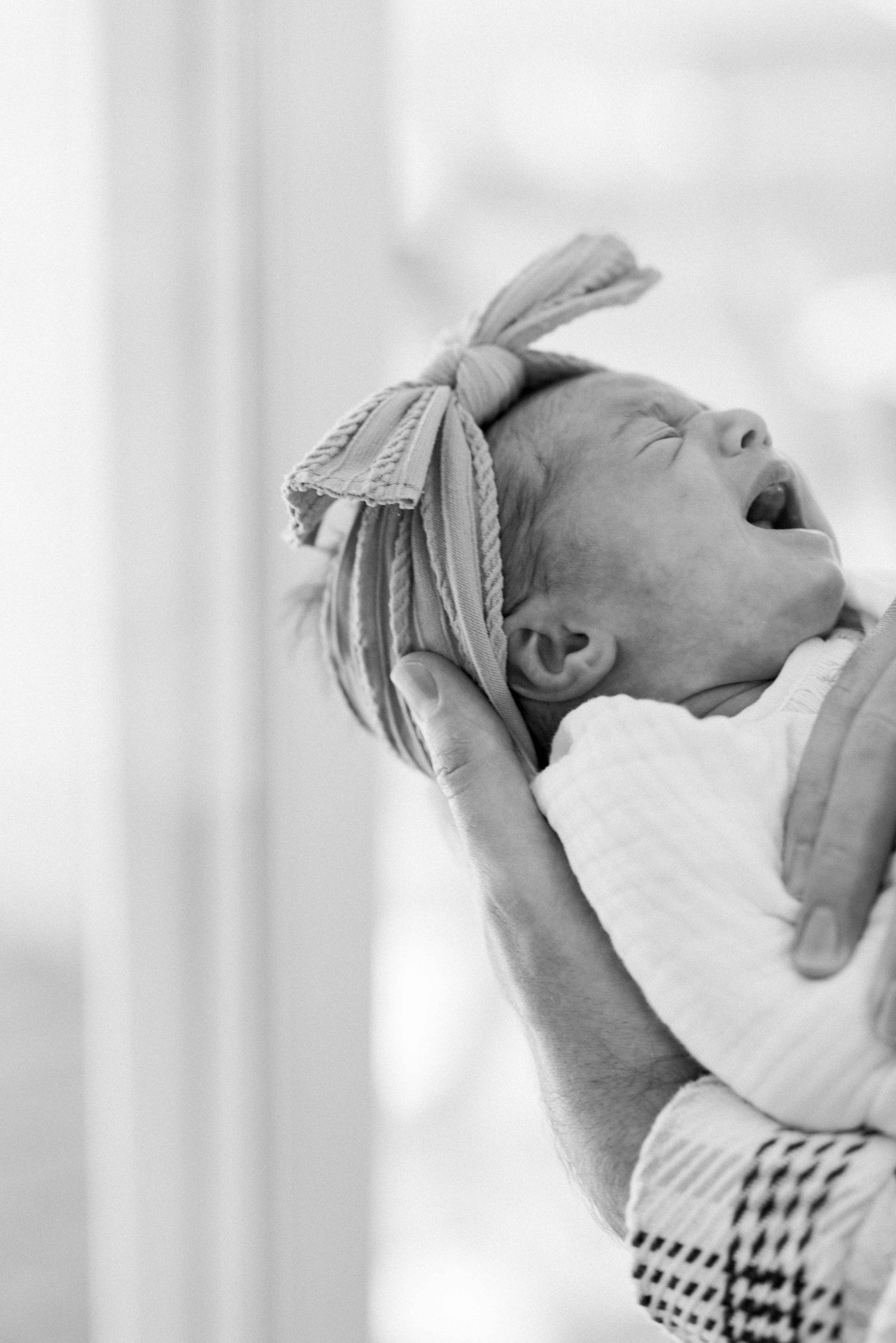 Close-up of crying newborn baby with bow headband being held up in black and white photography.