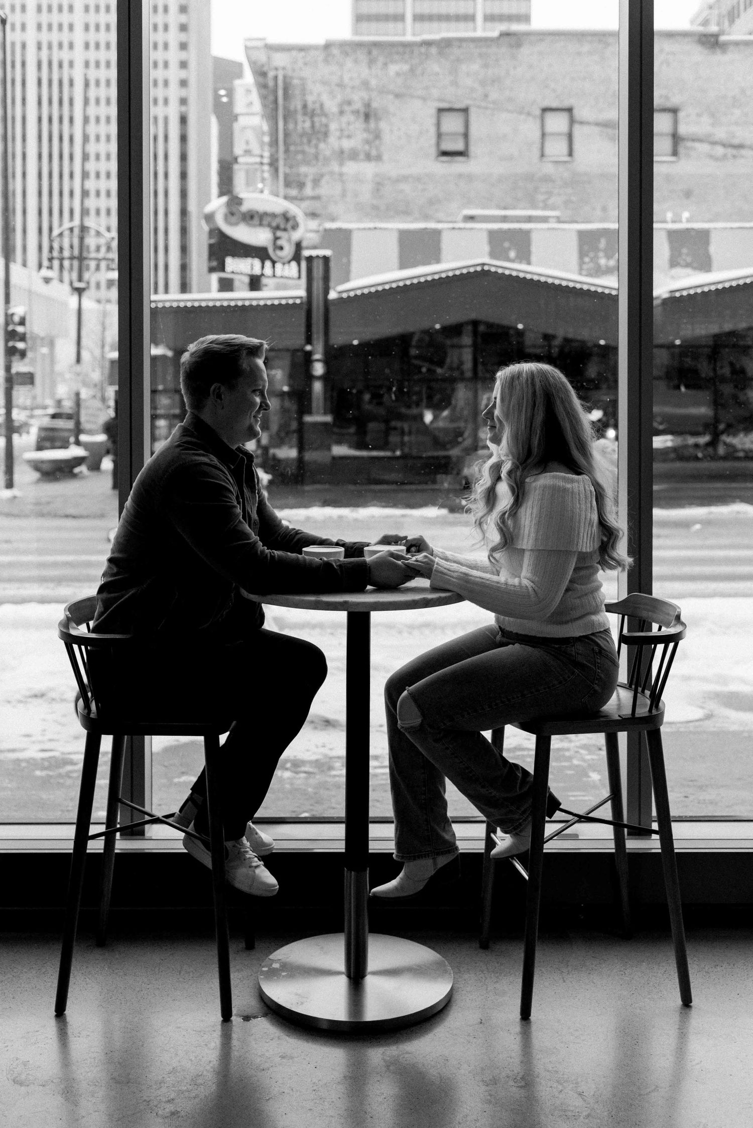 A silhouetted couple sits at a window-side cafe table in black and white.