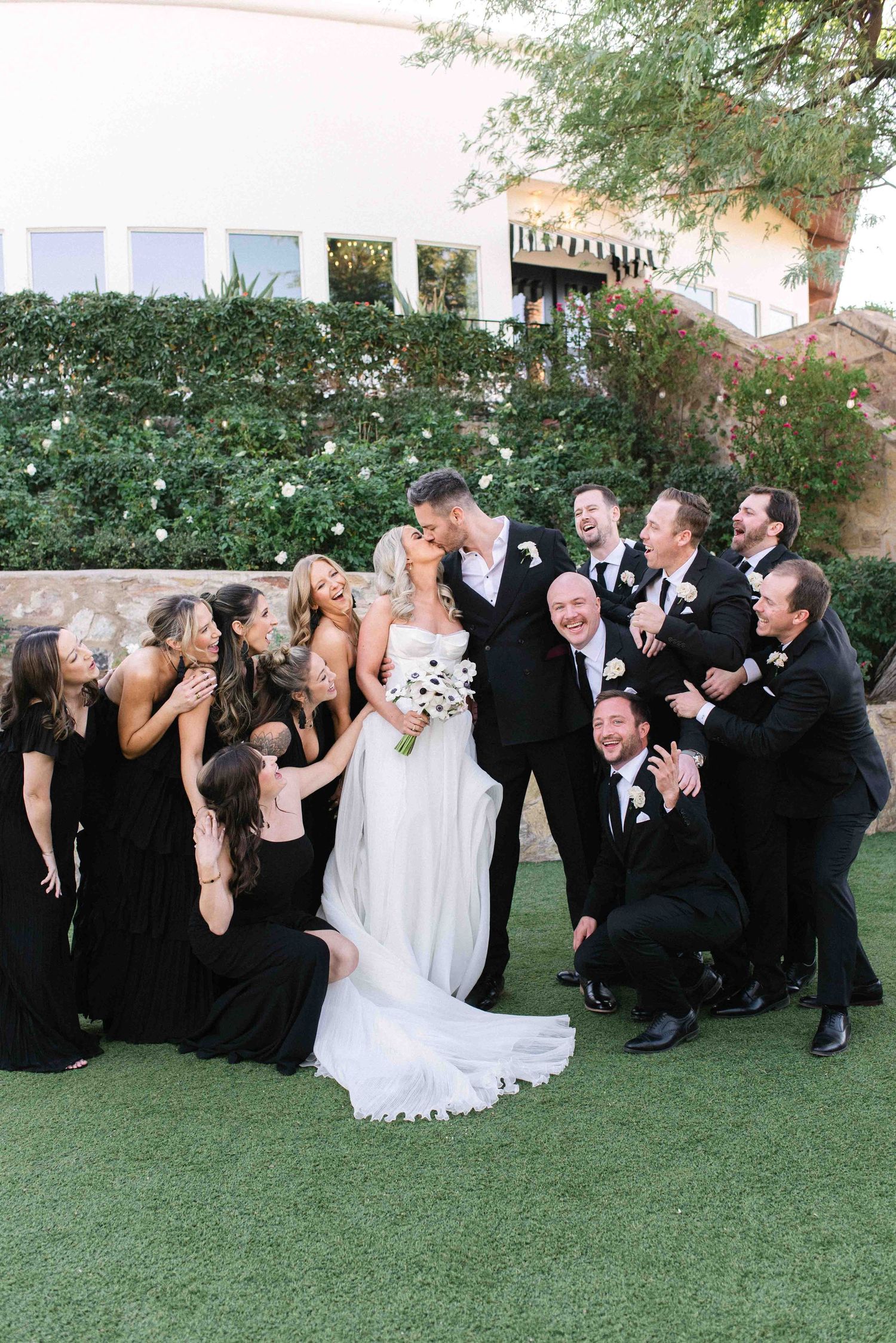 Wedding party in black attire gathers around bride for fun group photo on lawn.