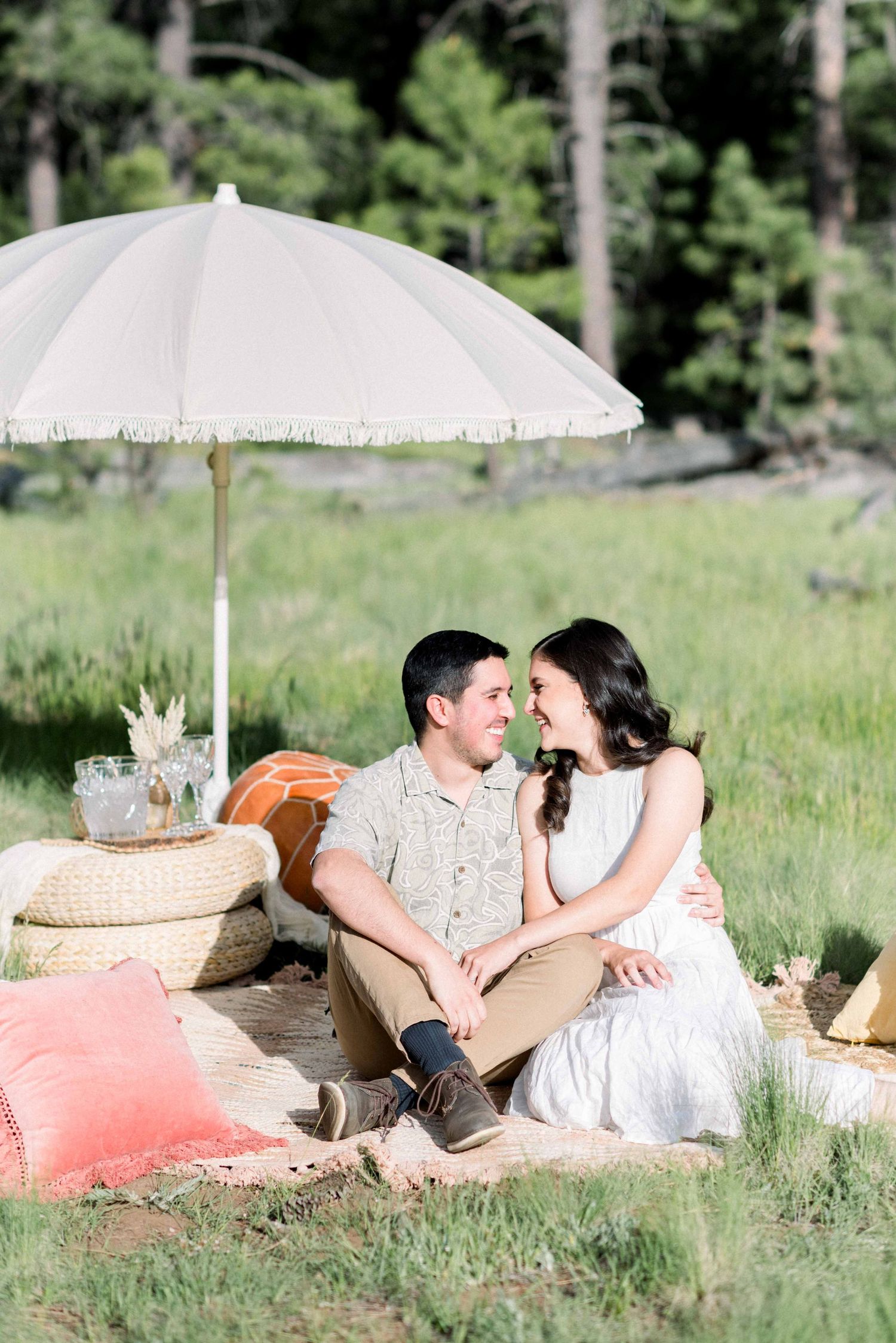 Couple sitting under white umbrella during romantic outdoor picnic photoshoot.