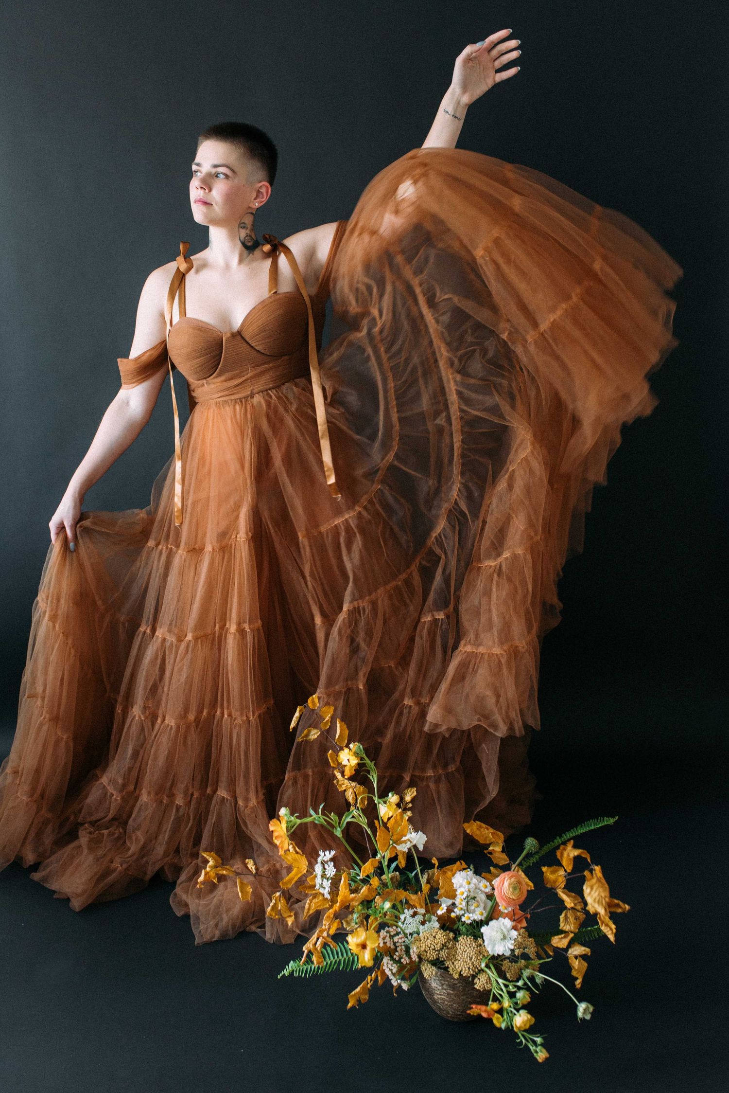 Dramatic pose in flowing brown tulle gown with floral arrangement against dark background.