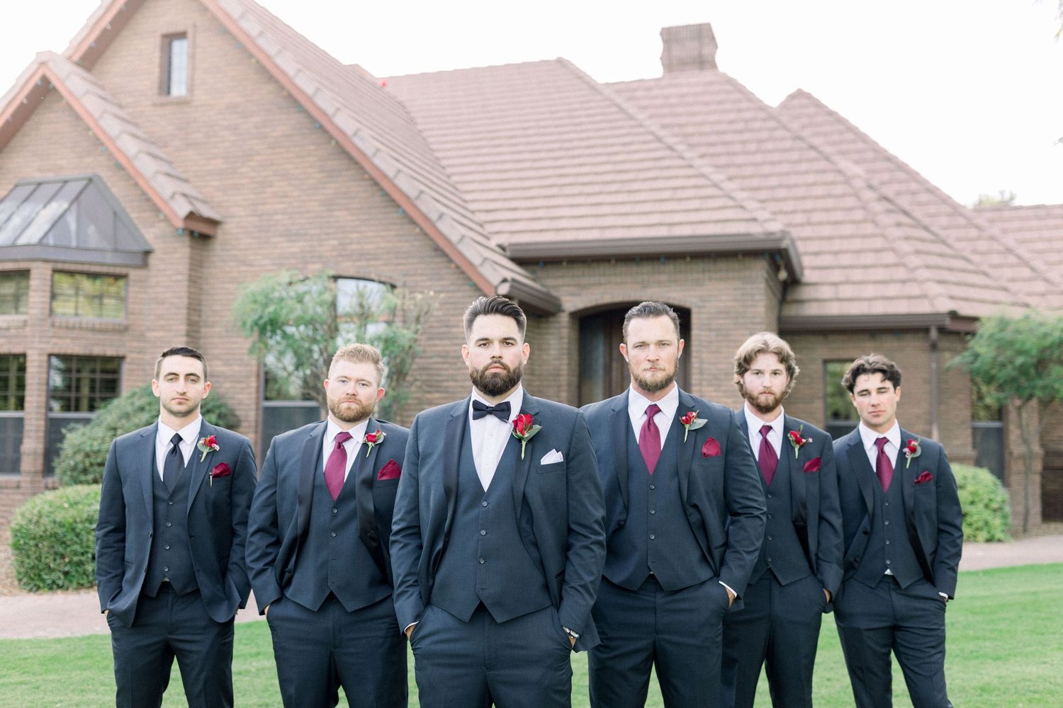 Wedding party of groomsmen in dark suits with red accents pose in front of a tan brick building.