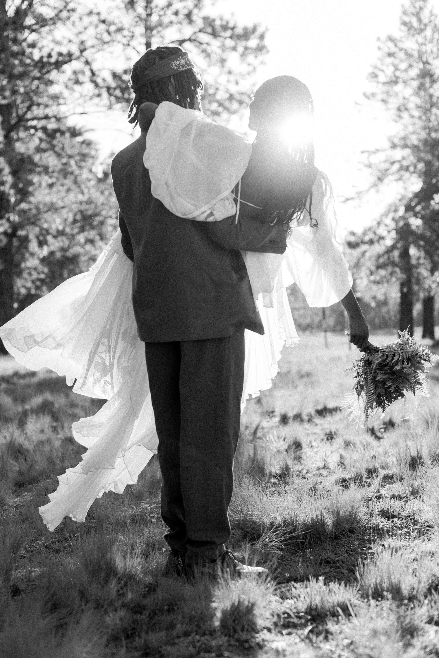 A romantic black and white wedding photo showing a groom carrying his bride uphill with sunlight streaming behind them.