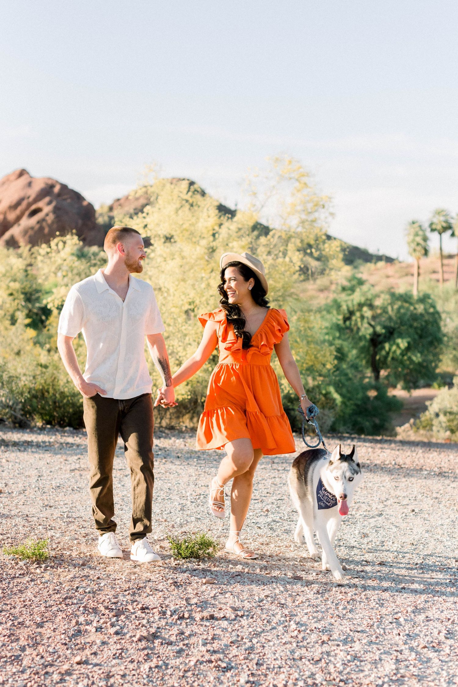 Couple walking with husky dog on desert trail with mountains in background.