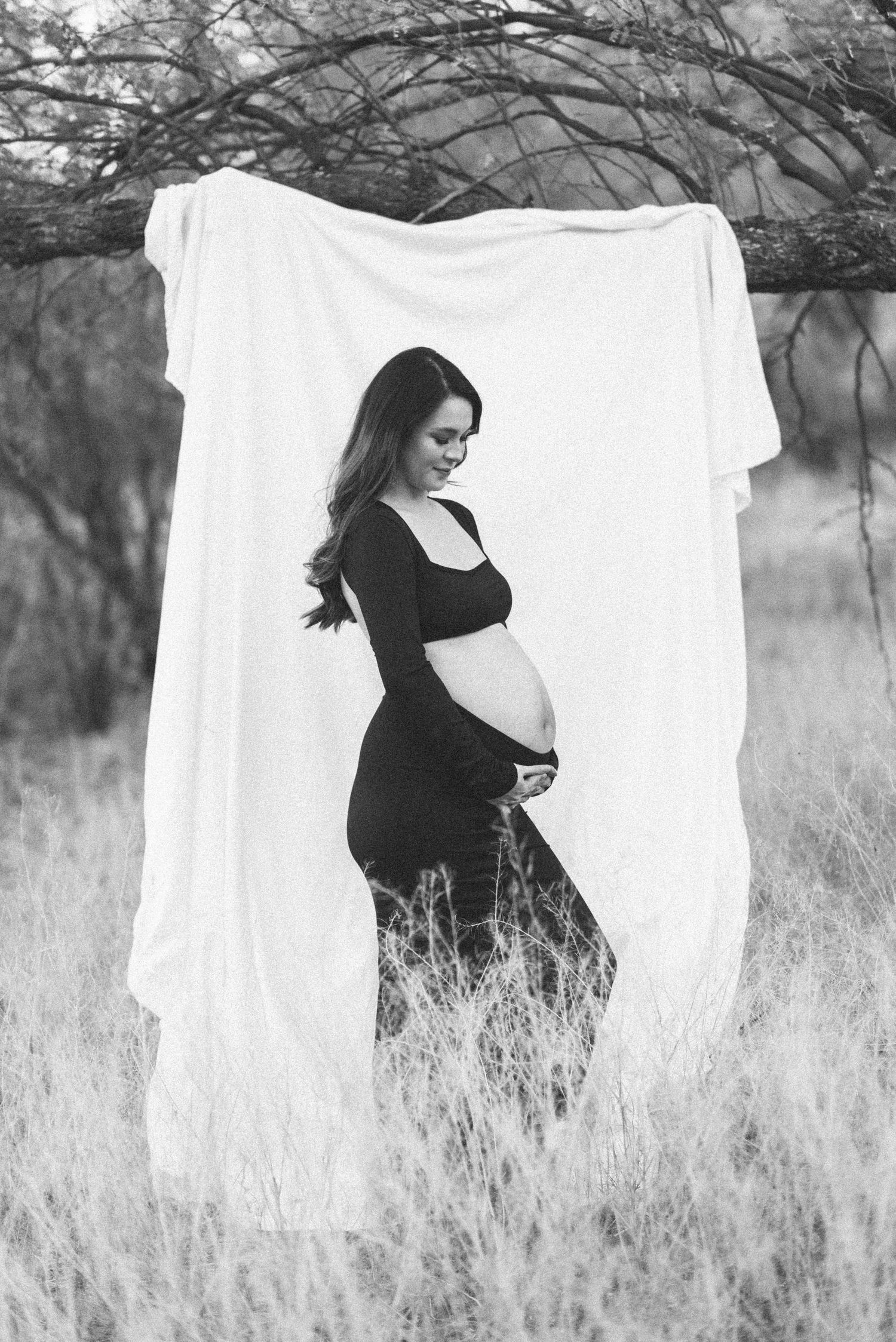 Black and white maternity photo against white backdrop in tall grass features expectant mother in elegant black dress.