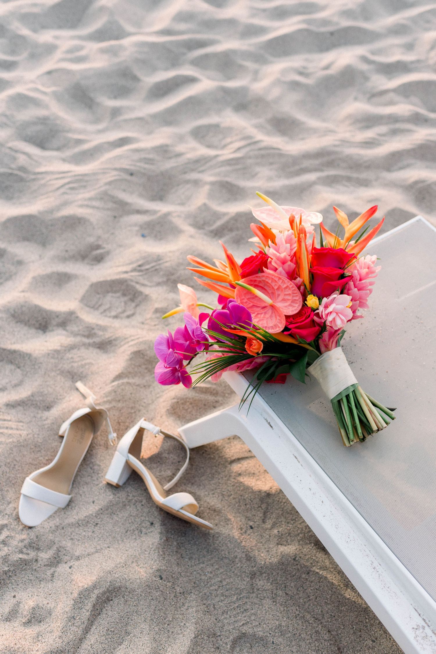 A vibrant tropical bridal bouquet with pink and orange flowers lies next to white sandals on beach sand.