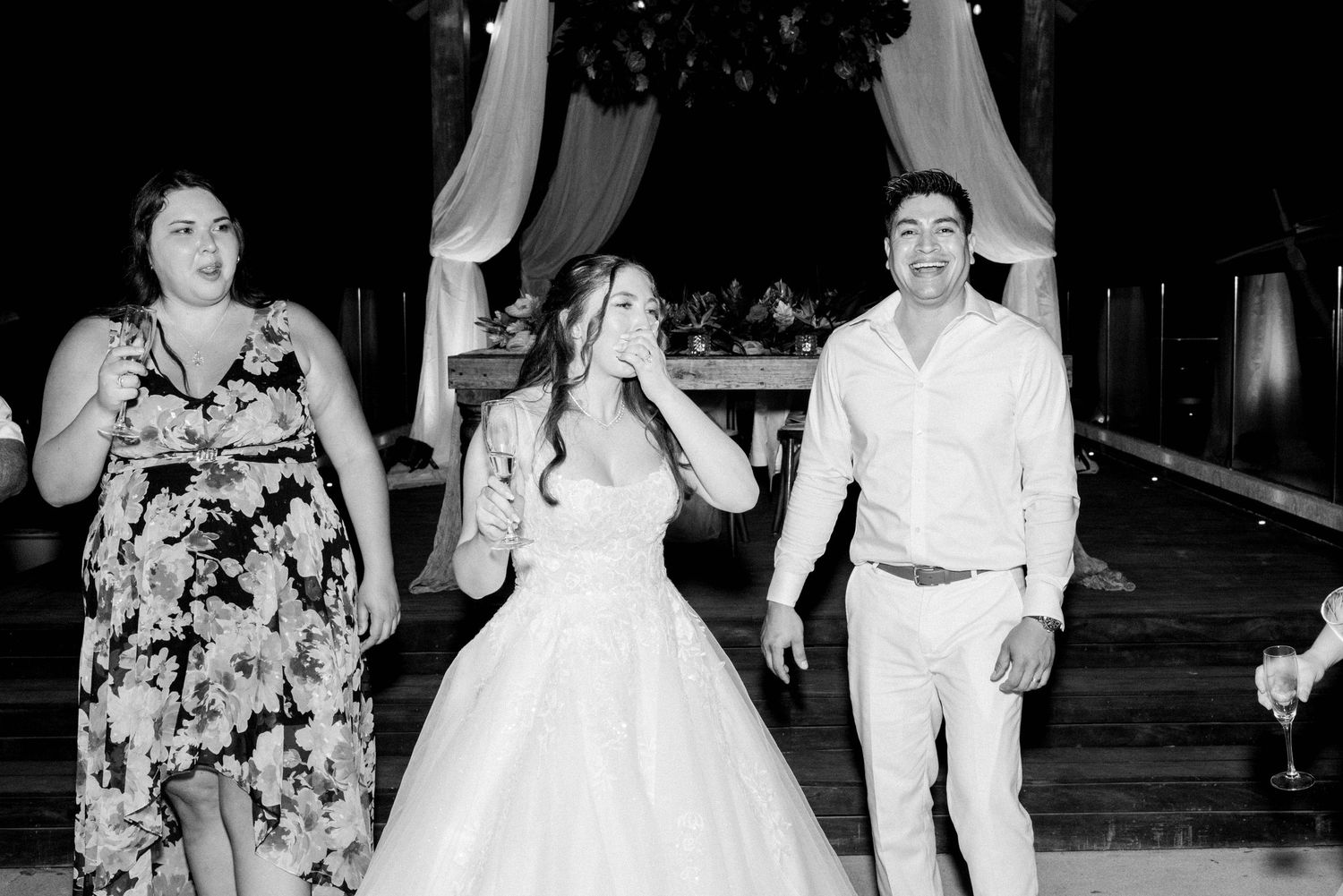 People celebrating with raised glasses beneath a wedding arch in black and white.