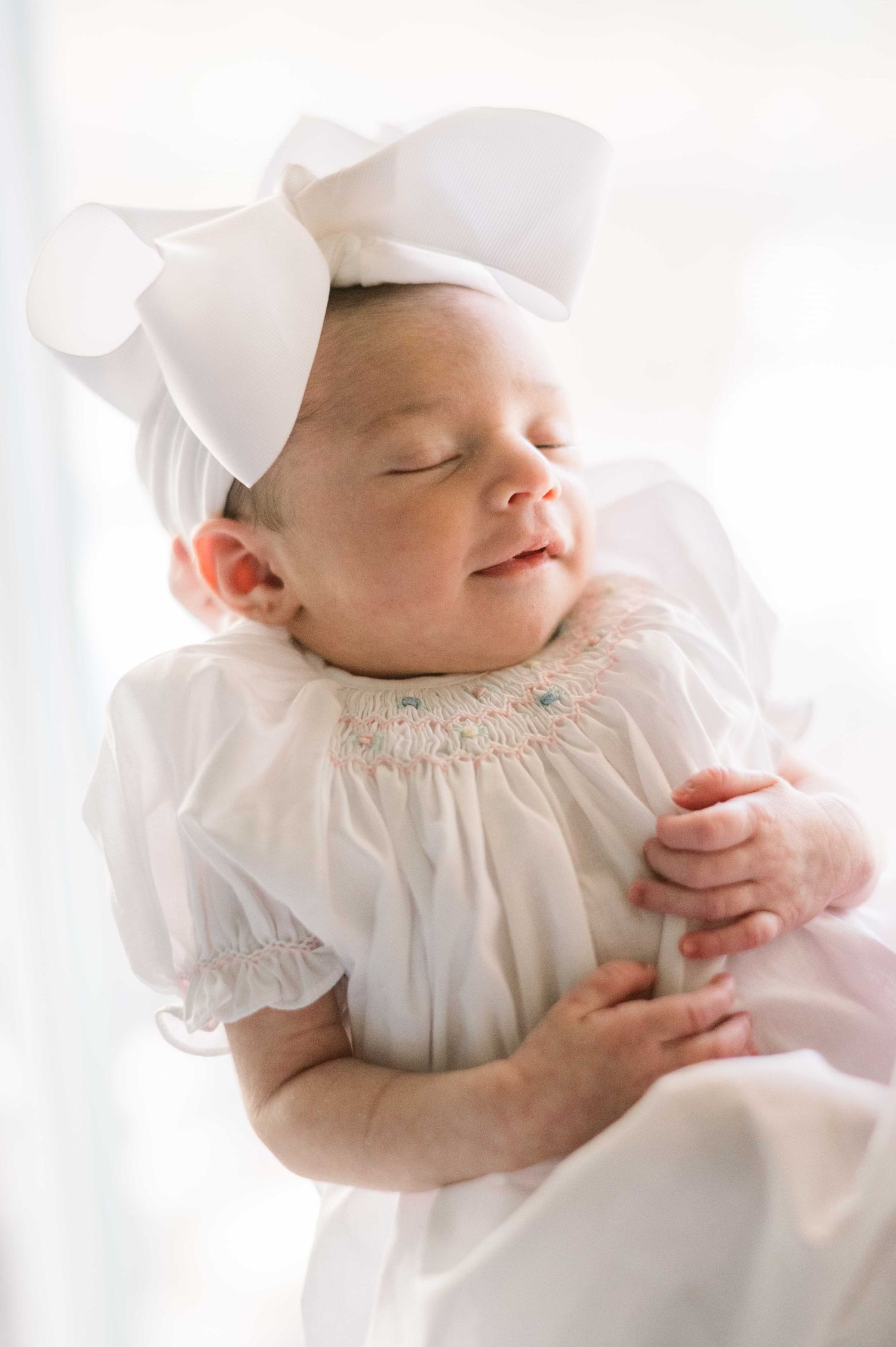 A sleeping baby in white smocked dress and large bow rests peacefully.