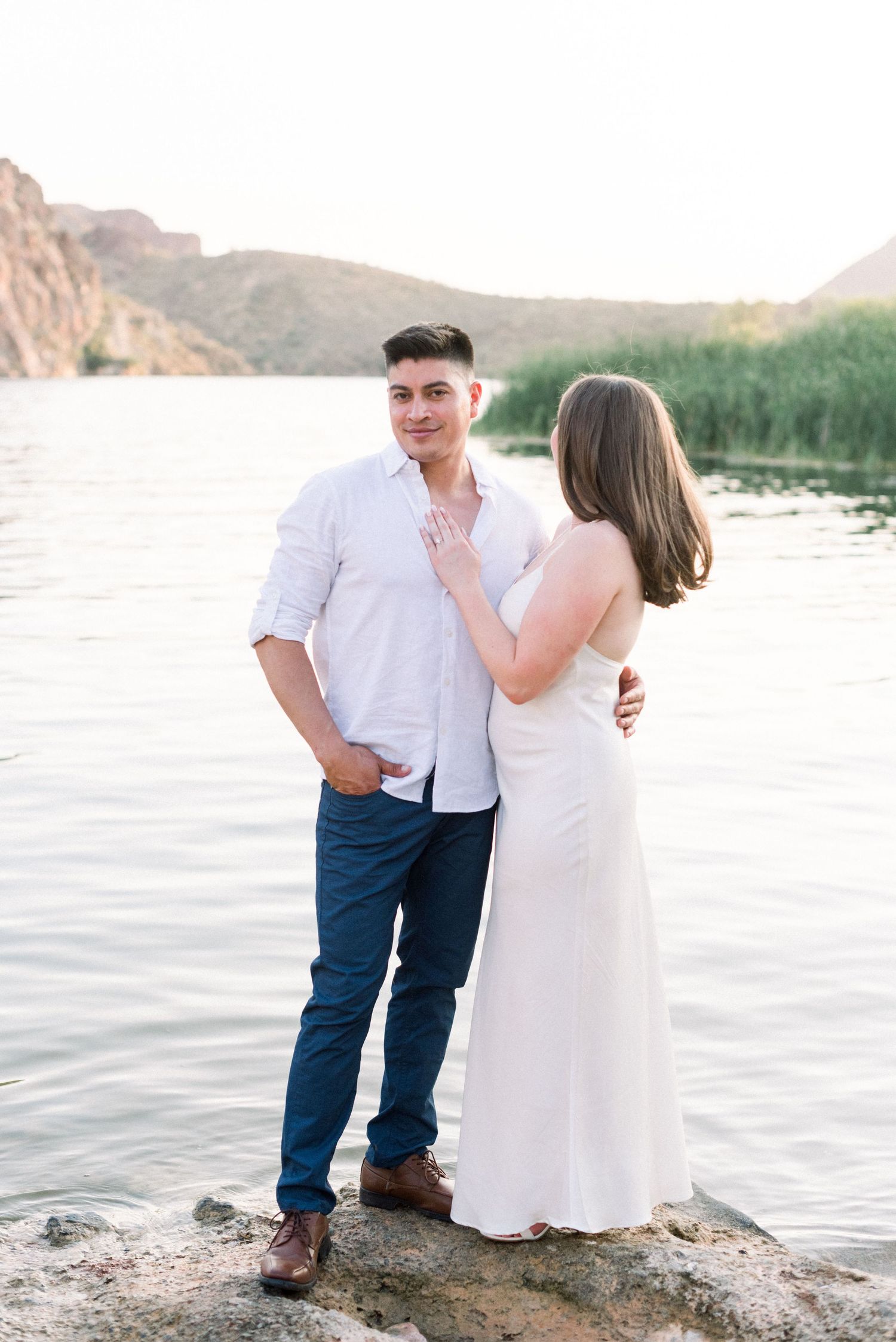 A couple stands on rocks by a lake with mountains and sunset in the background.