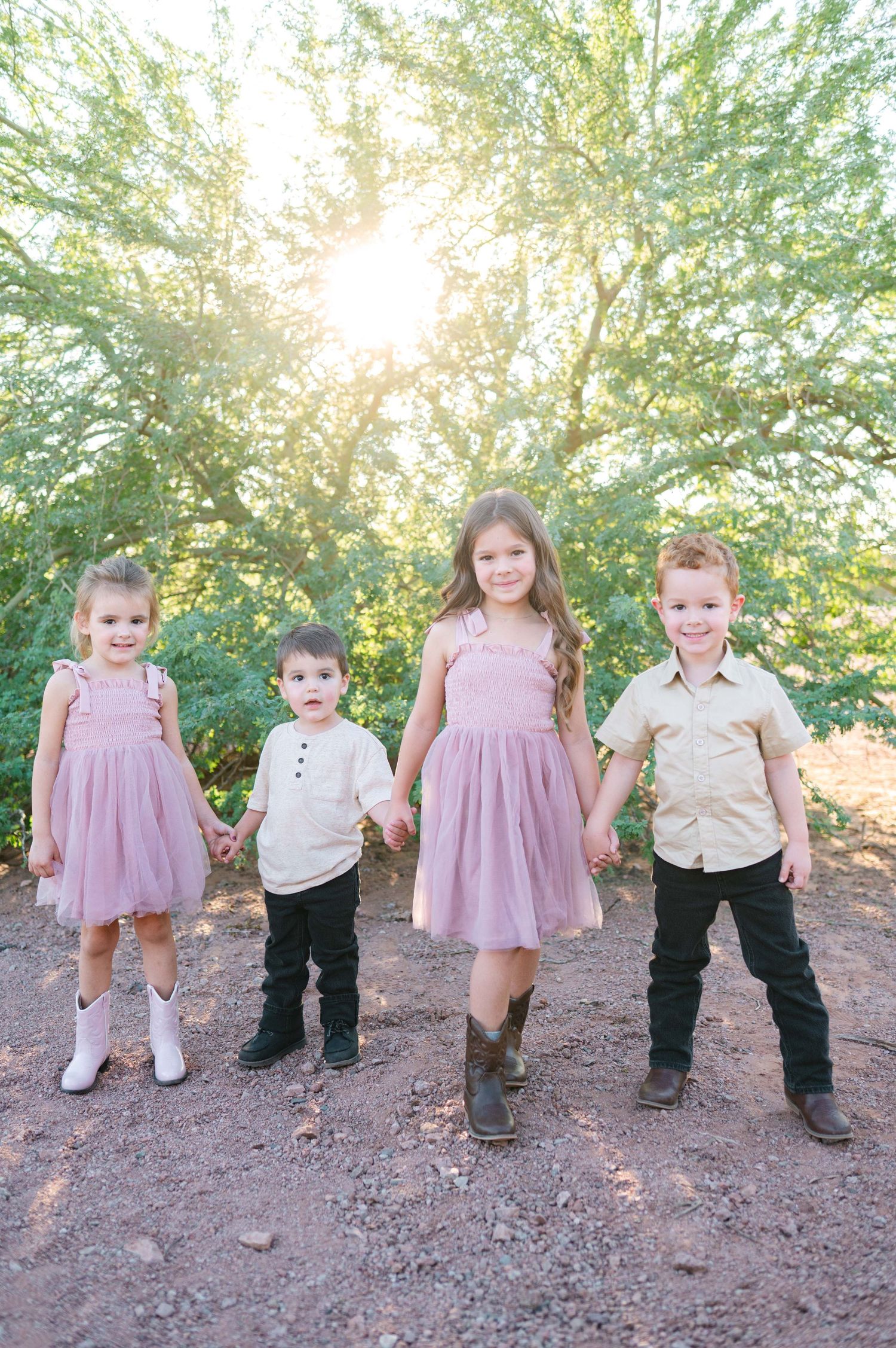 Children in pink dresses and khaki outfits walk together on a dirt path under sunlit trees in a sweet outdoor photo series.