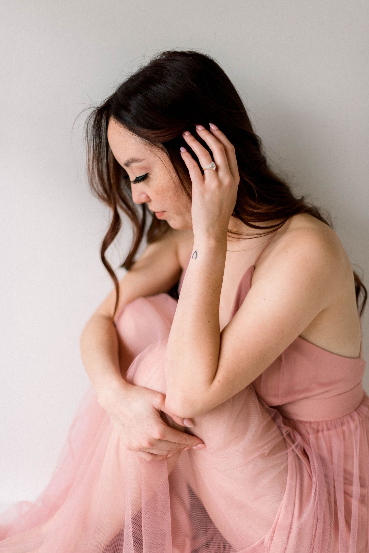 Figure in pink tulle gown leans forward with hand in dark hair against white backdrop.
