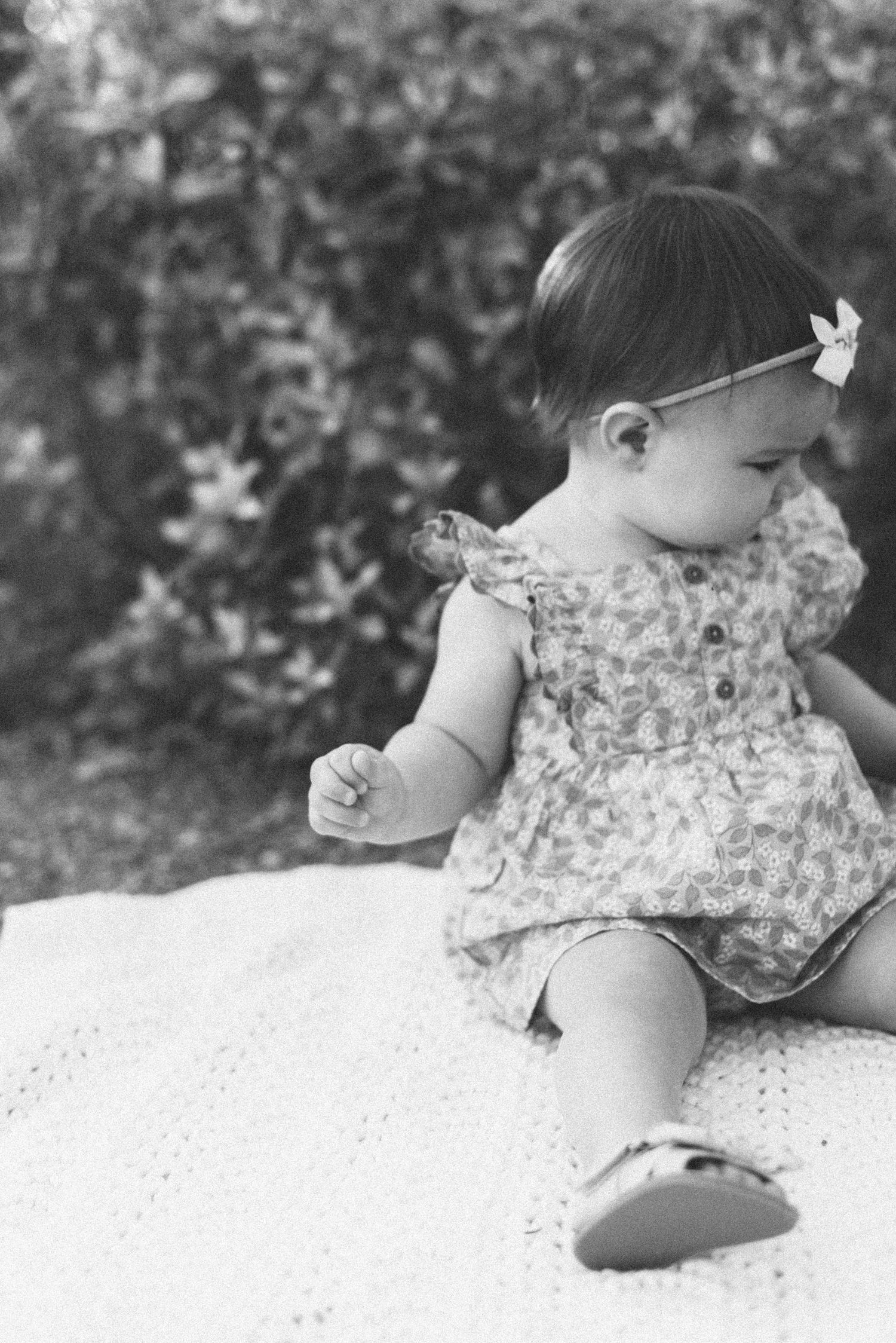 Vintage black and white photo of a baby in floral dress sitting on white blanket.