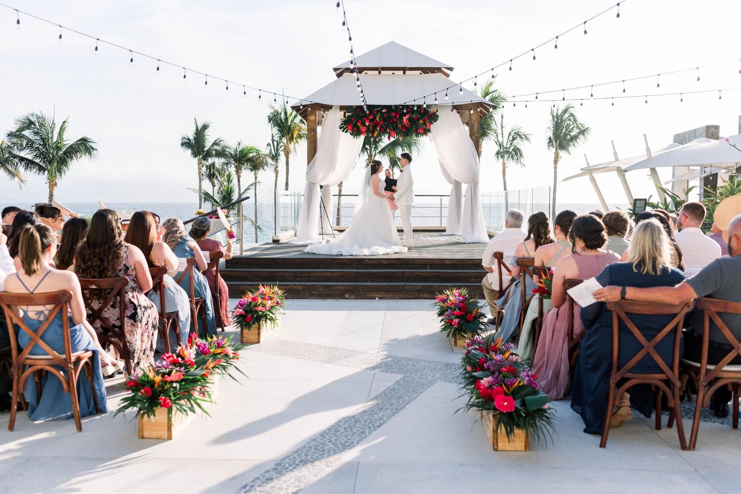 Outdoor wedding ceremony setup with white gazebo and cross-back chairs lined with floral arrangements.