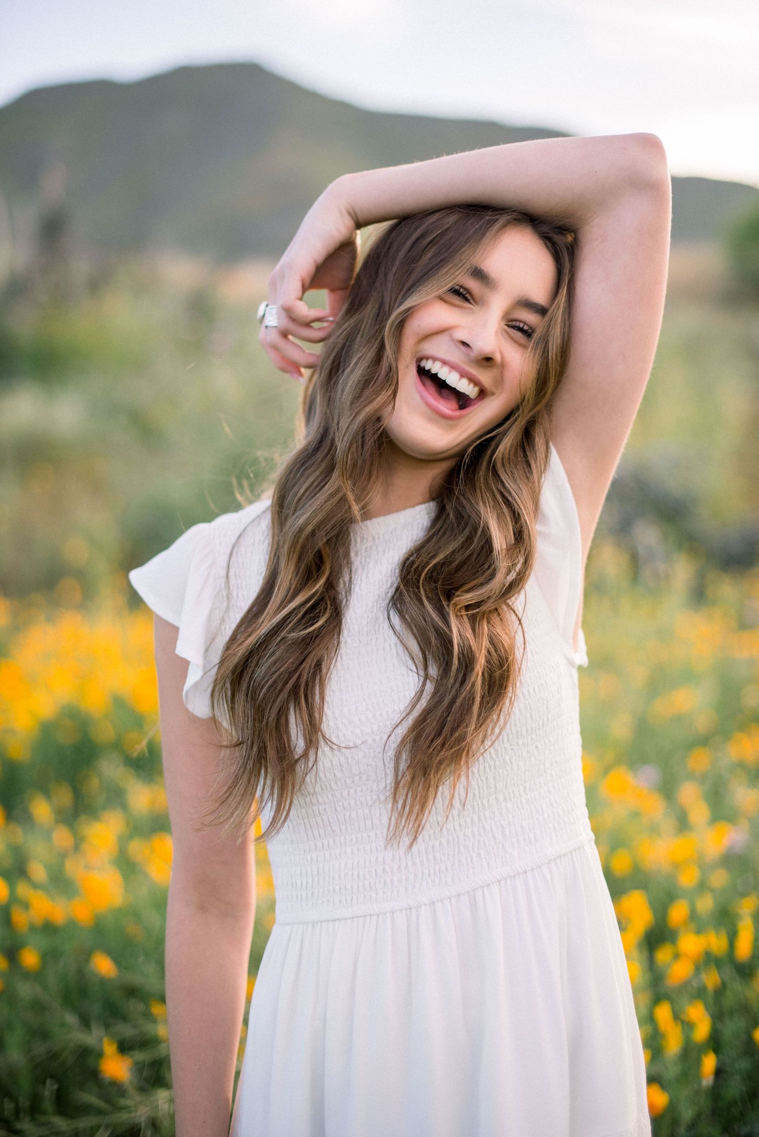 Joyful pose in white dress surrounded by yellow wildflowers and mountain scenery.