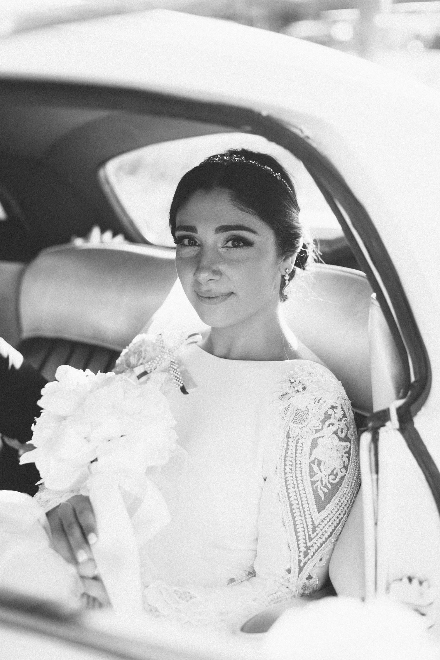 A bride in an elegant lace wedding dress sits in a car, captured in black and white.