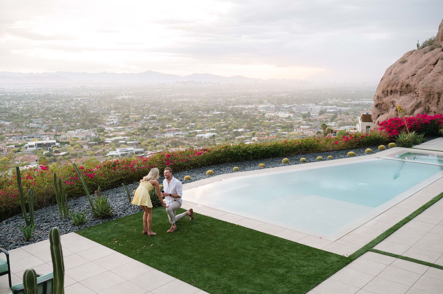 A couple stands by an infinity pool overlooking a scenic cityscape at sunset.