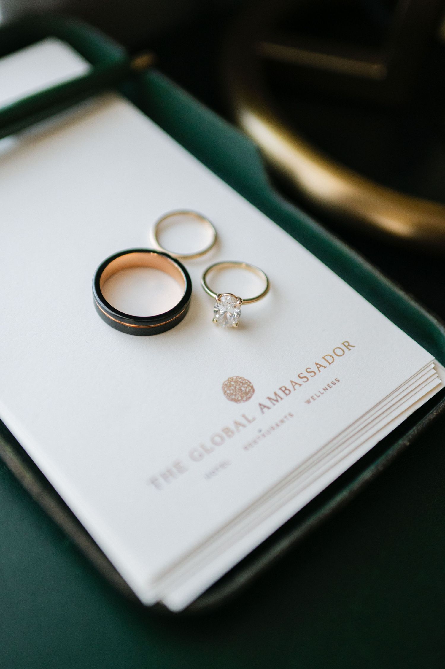 Wedding rings rest elegantly on white stationery with gold embossed text against a dark green background.