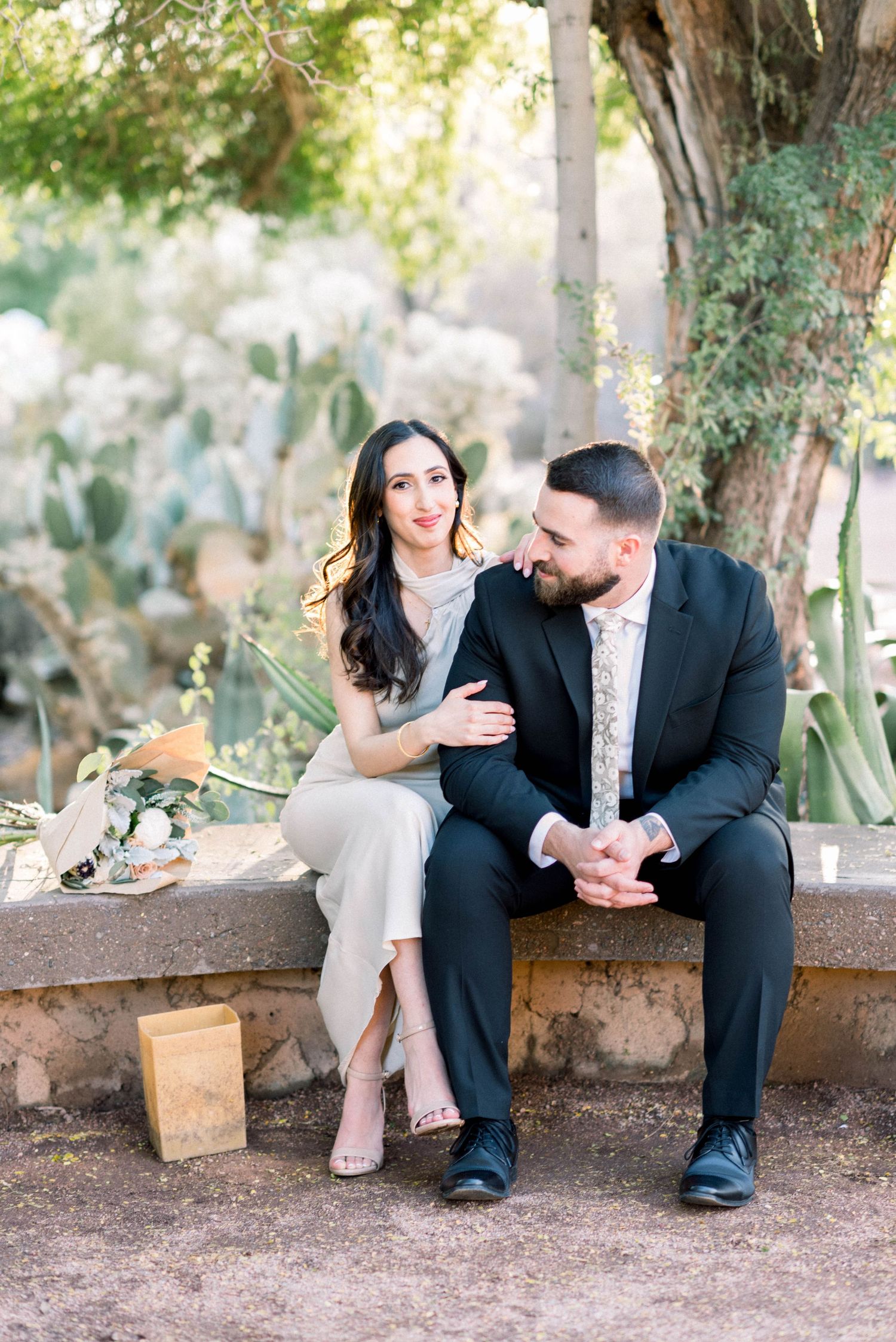 A couple in formal attire sits together on a rustic stone bench in a garden setting during an engagement photoshoot.