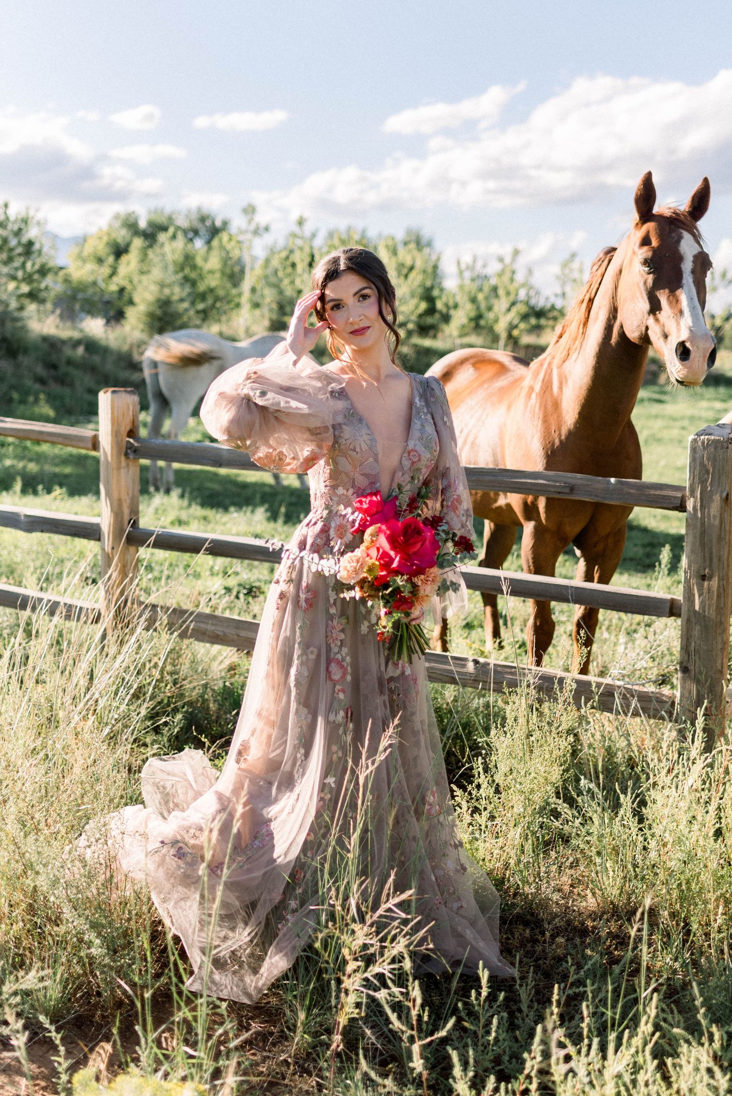 Woman in flowy dress poses near wooden fence with horses grazing in summer field.