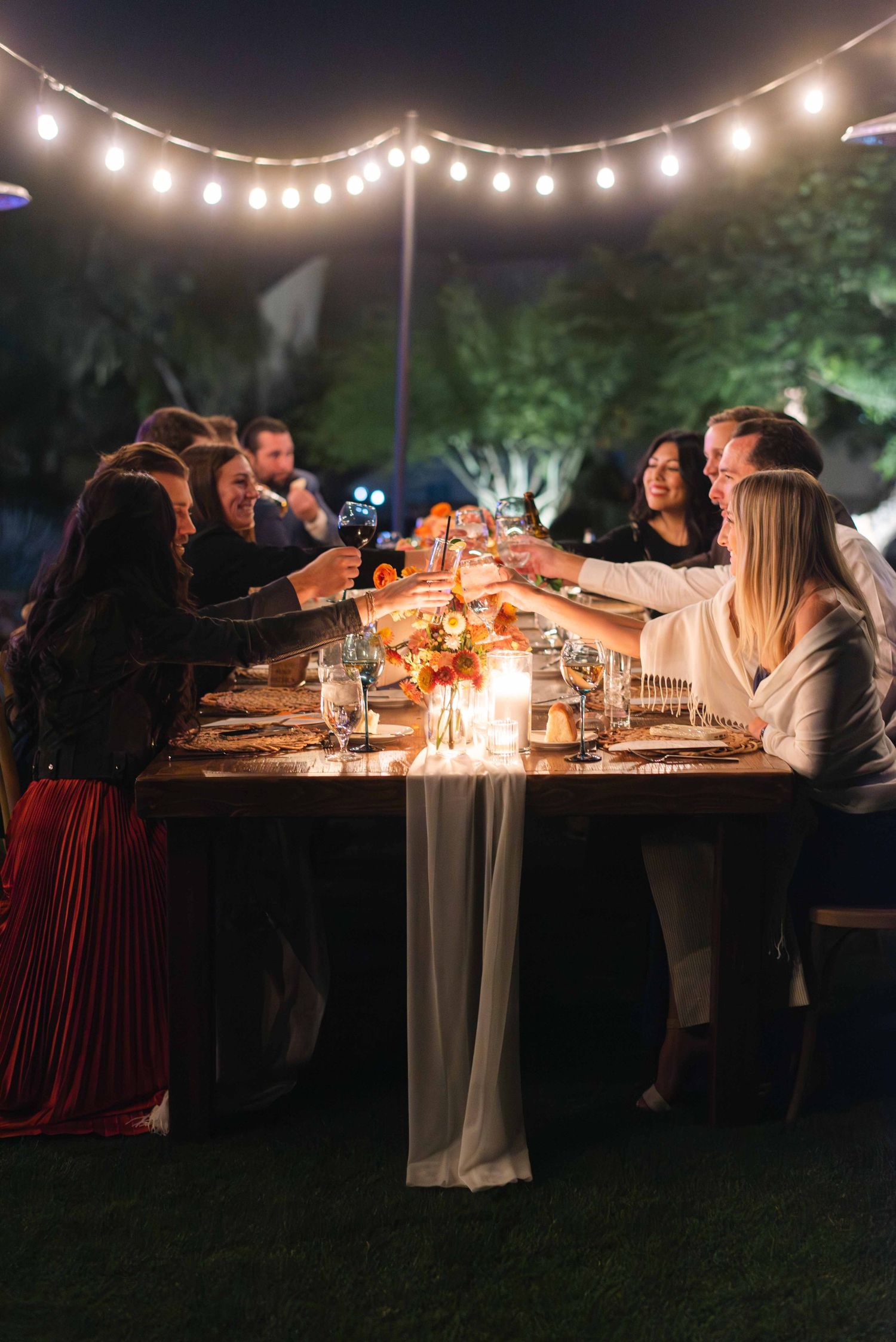 Outdoor evening dinner party under string lights with guests gathered at a long table illuminated by candlelight.