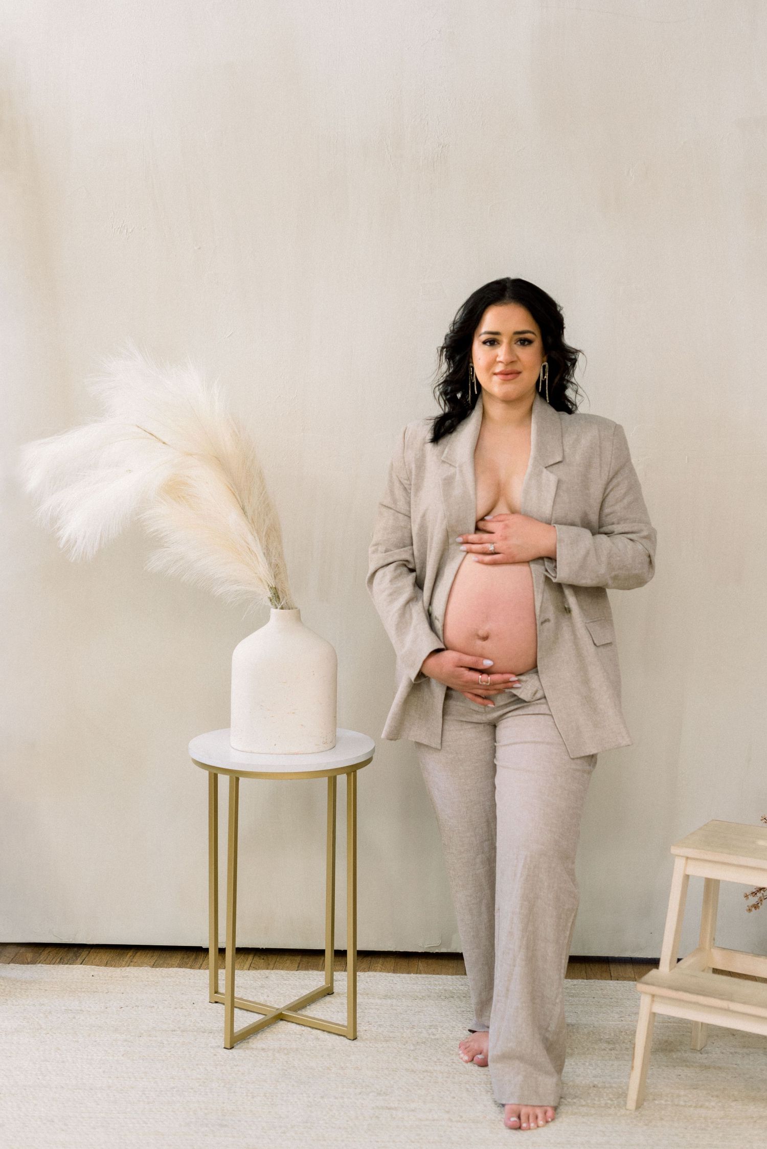 Maternity portrait in soft beige pantsuit with pampas grass decor on modern gold accent table.