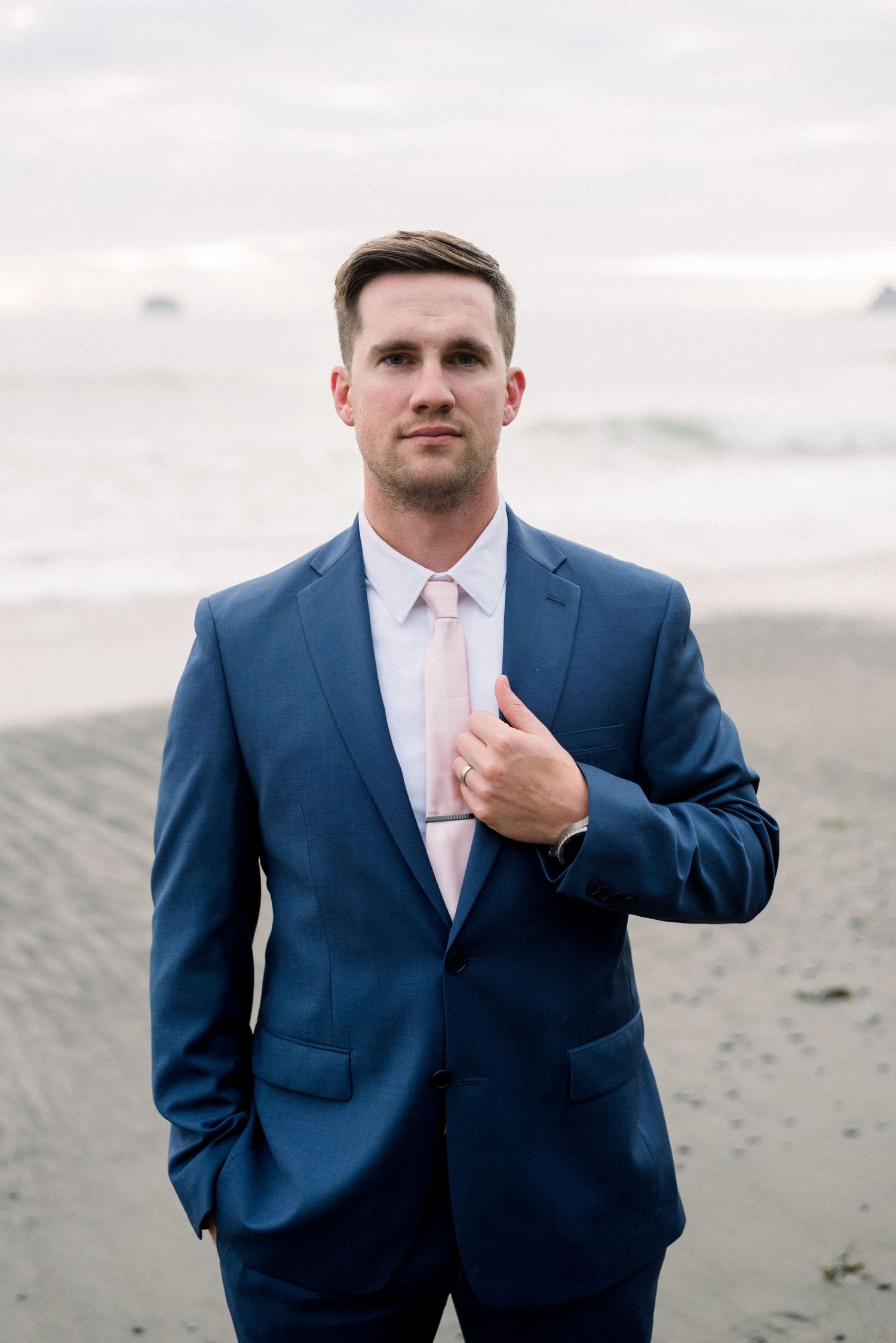 Person wearing navy suit and pink tie poses on beach at sunset.