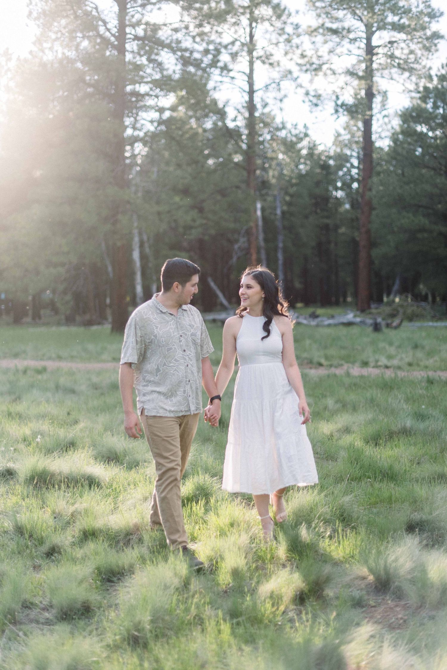 A couple walks hand in hand through a sun-drenched forest clearing wearing coordinated casual outfits.